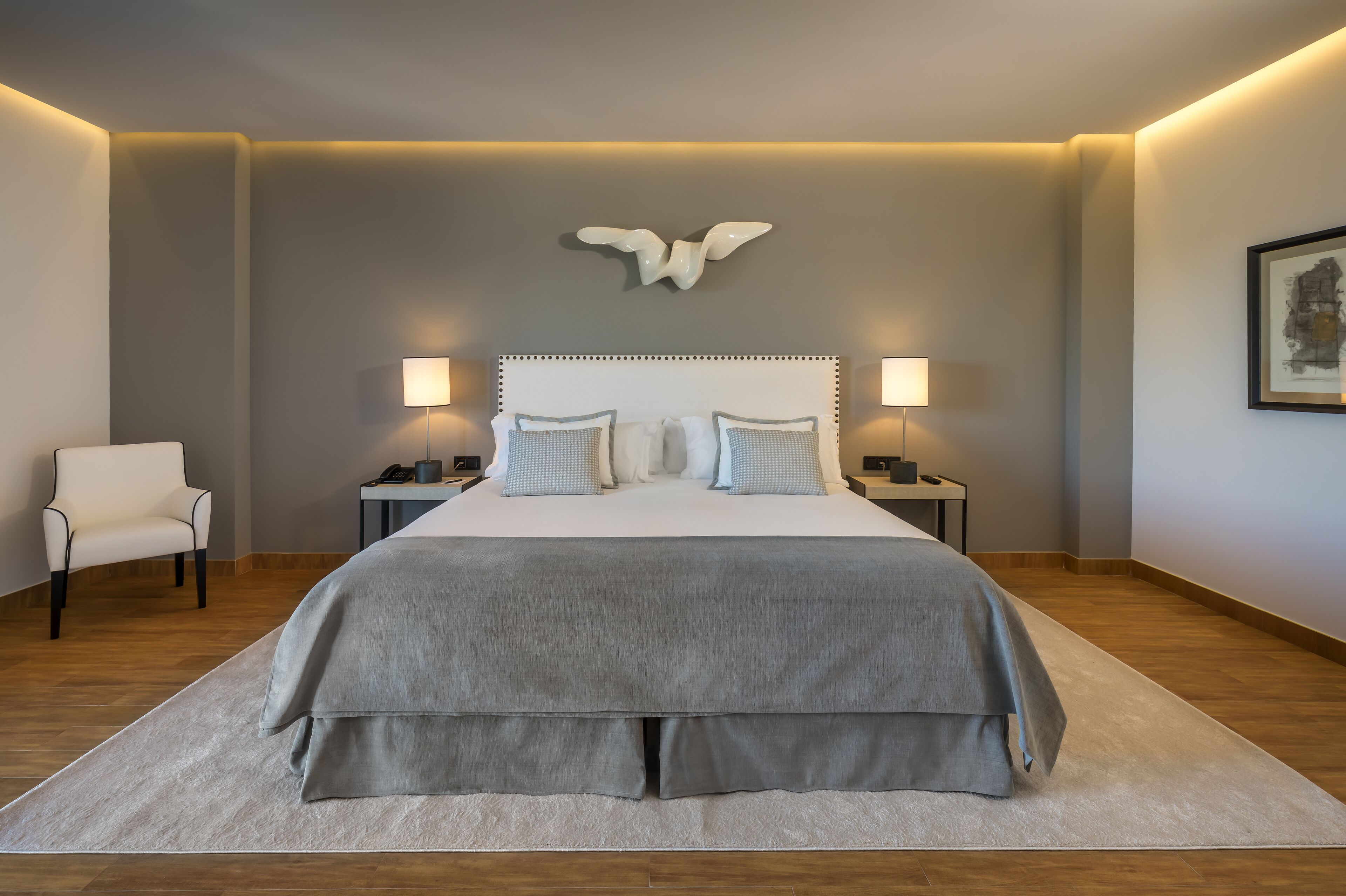 Une chambre moderne avec un grand lit, deux tables de chevet, une chaise et un luminaire fixé au mur.