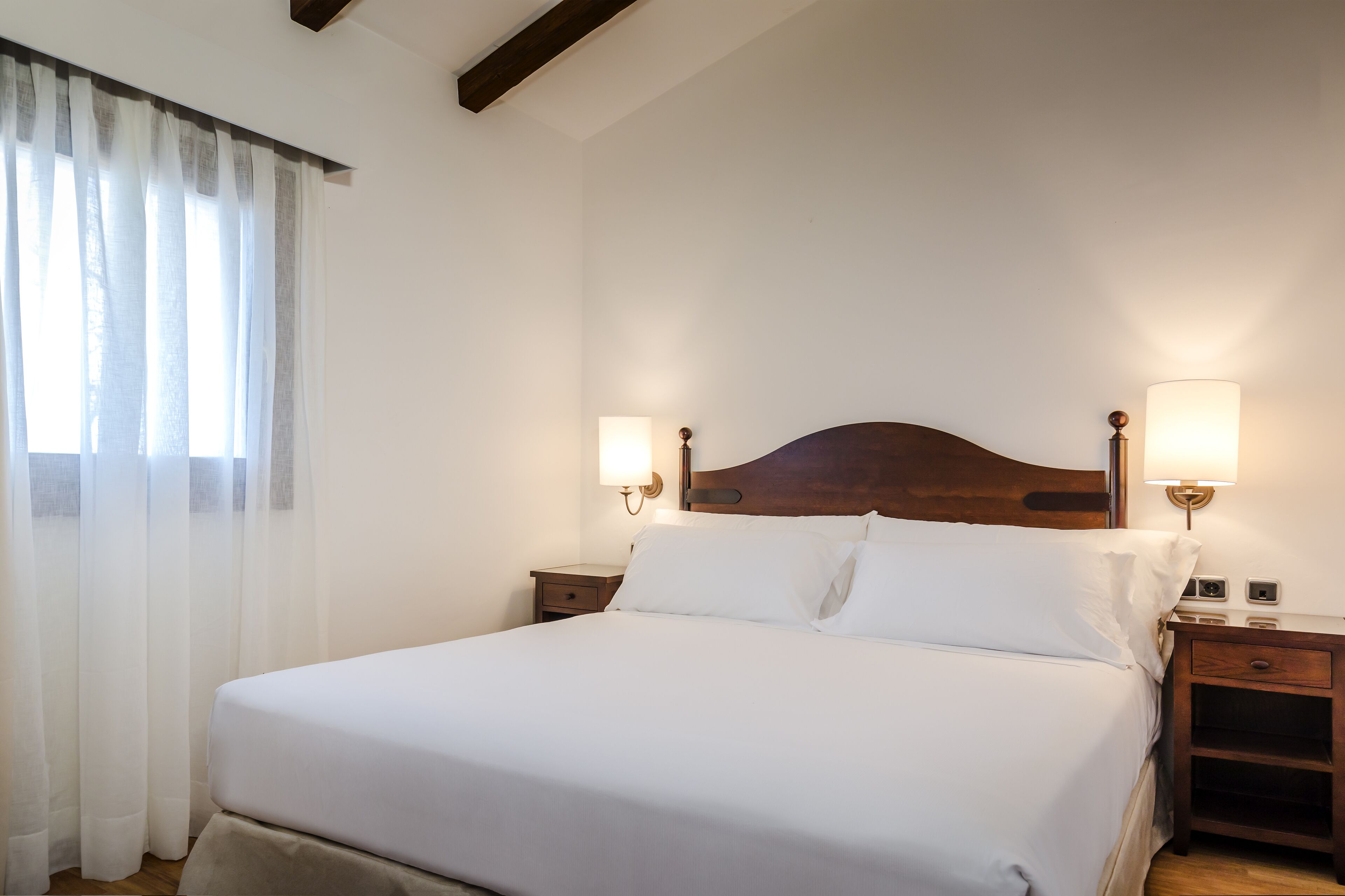 A neatly made bed with white linens, two bedside tables with lamps, and sheer curtains on a window.