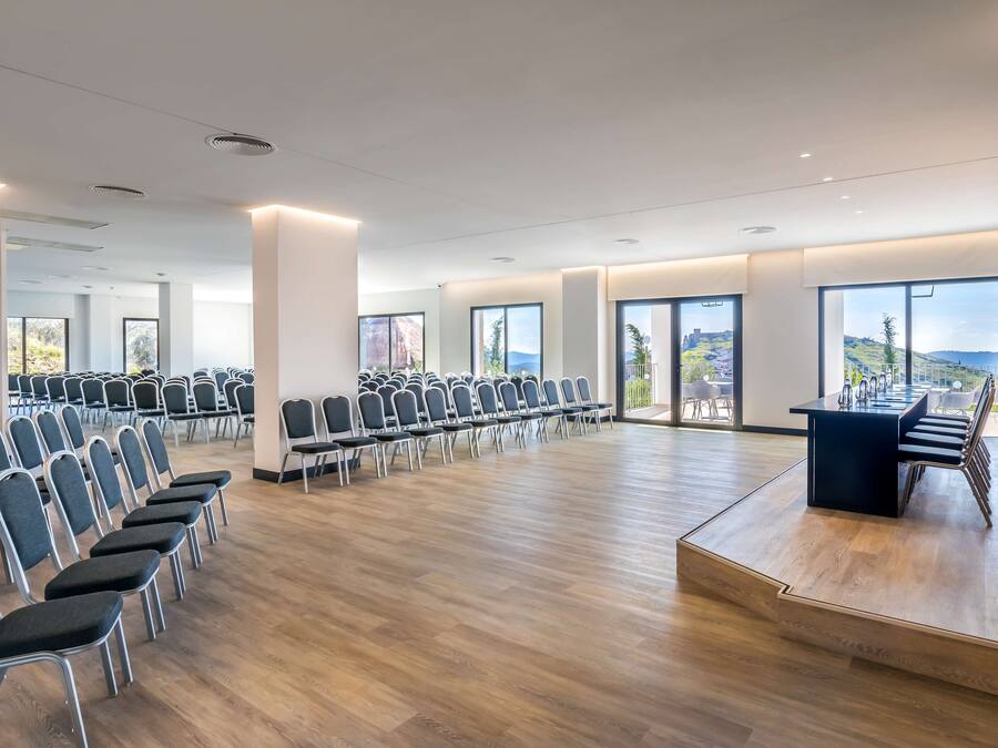 Una amplia sala de conferencias con filas de sillas y un atril, grandes ventanales que ofrecen vistas al exterior.