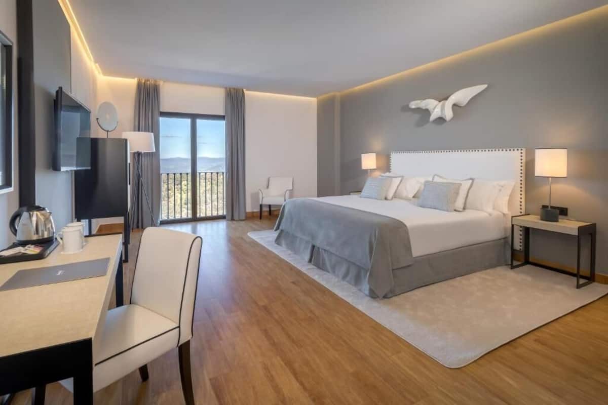 A modern hotel room with a large bed, a desk with a chair, a TV, and a balcony with a view.