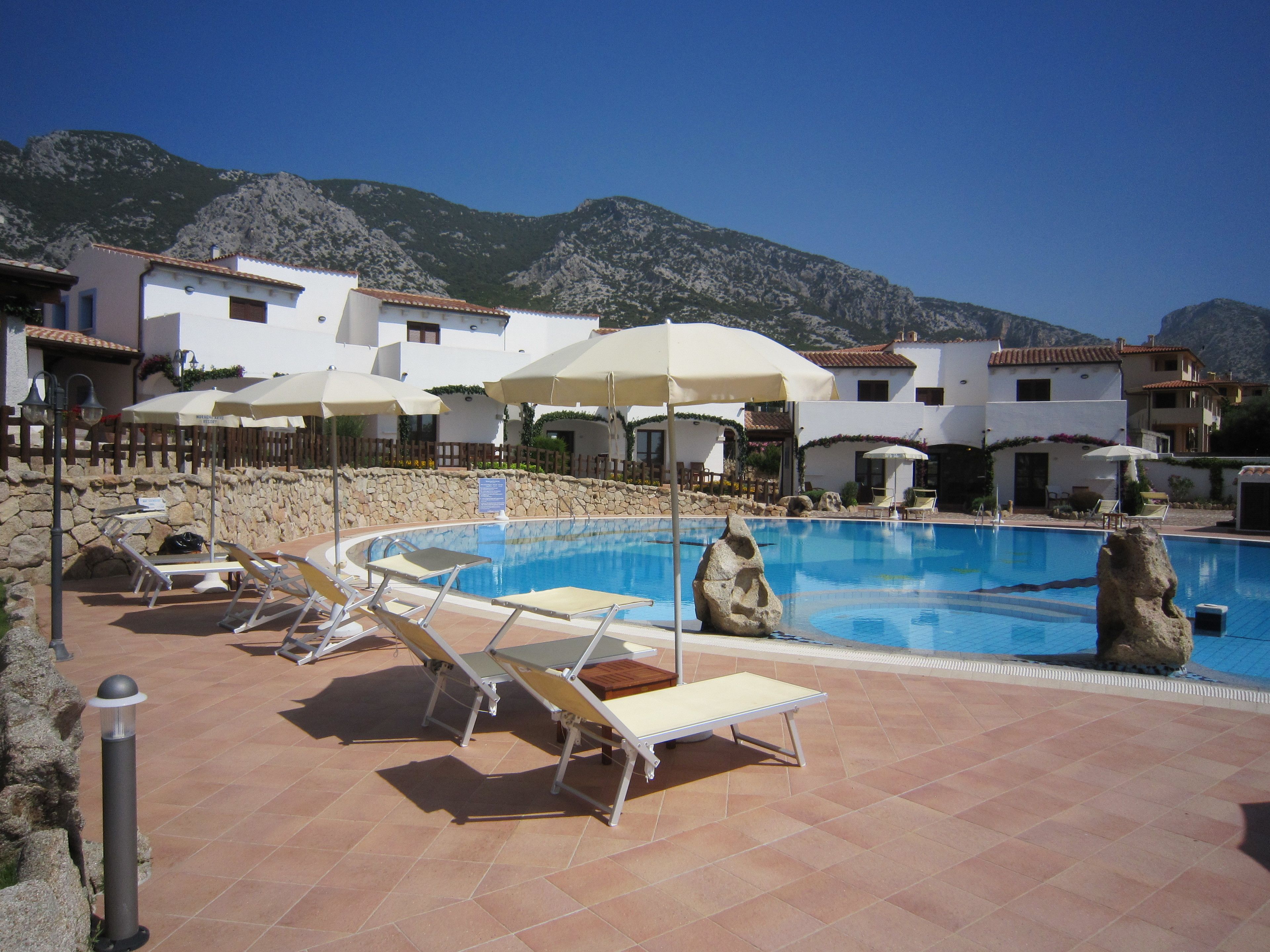 A pool area with lounge chairs and umbrellas, surrounded by buildings with white walls and red-tiled roofs, set against a mountainous backdrop.