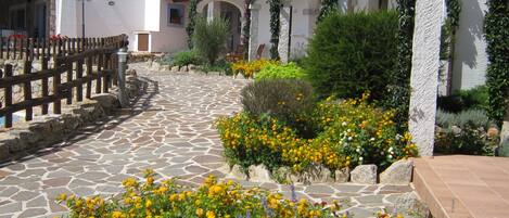 Un sentier pavé de pierres mène à un bâtiment blanc, entouré d’une clôture en bois et agrémenté de parterres de fleurs jaunes et violettes.