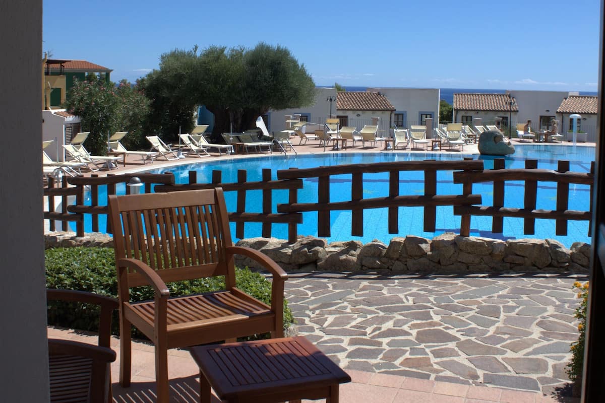 Un área junto a la piscina con una silla y mesa de madera, un patio con baldosas de piedra, y vistas a una piscina con tumbonas y edificios al fondo.