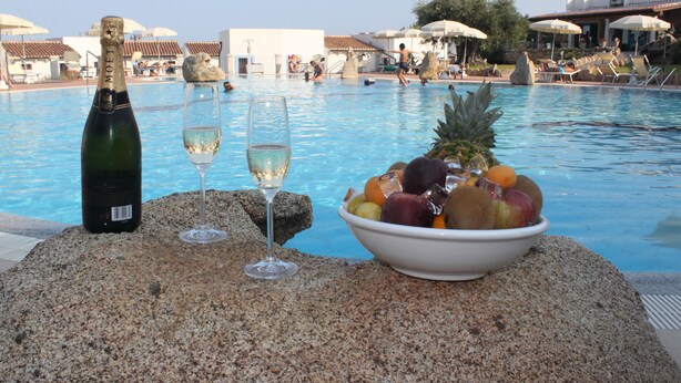 A poolside setting with a bottle of champagne, two glasses, and a bowl of fruit. - Hotel Nuraghe Arvu (Dorgali)