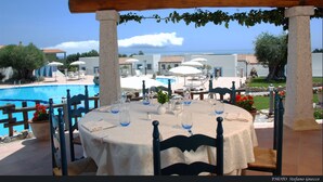 An outdoor dining area with tables set for two, overlooking a swimming pool and a view of the sea. - Hotel Nuraghe Arvu (Dorgali)