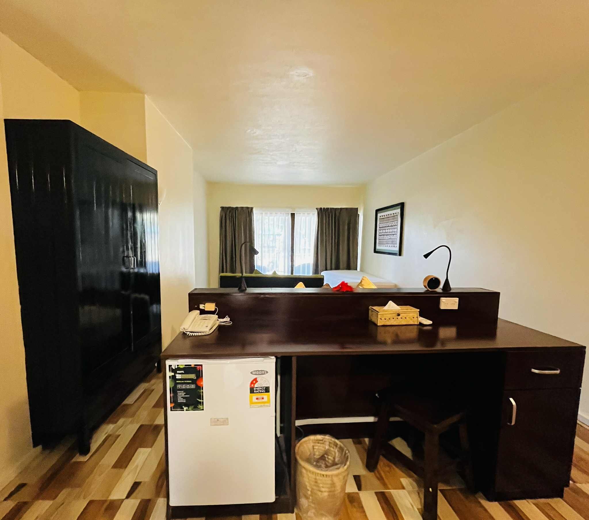A compact hotel room with a kitchenette, a small refrigerator, a desk with a chair, a trash bin, and a window with curtains.