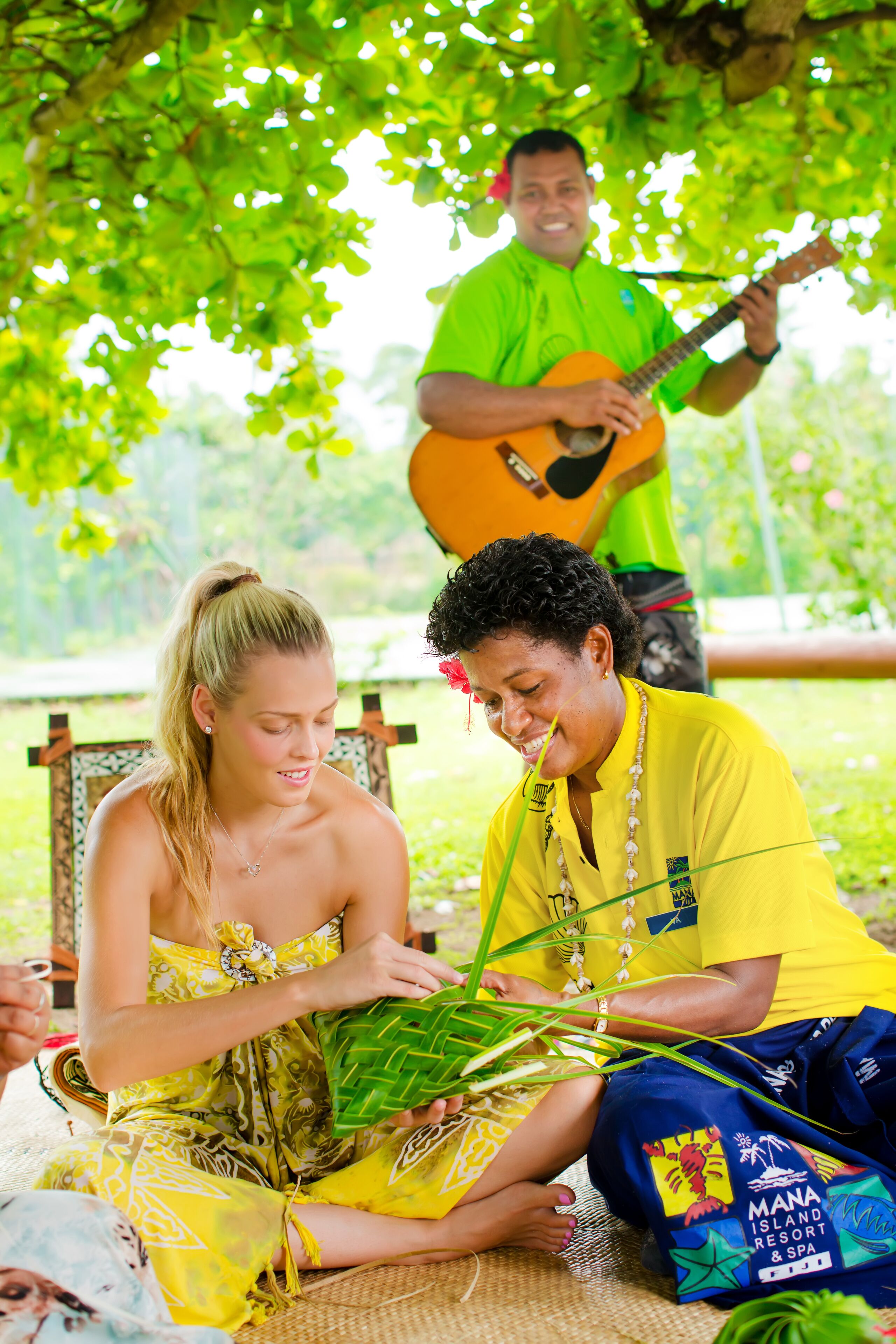 Una persona sentada sobre una estera tejida bajo un árbol, otra persona de pie con una guitarra, y una bolsa con la inscripción “MANA Island Resort & Spa”.