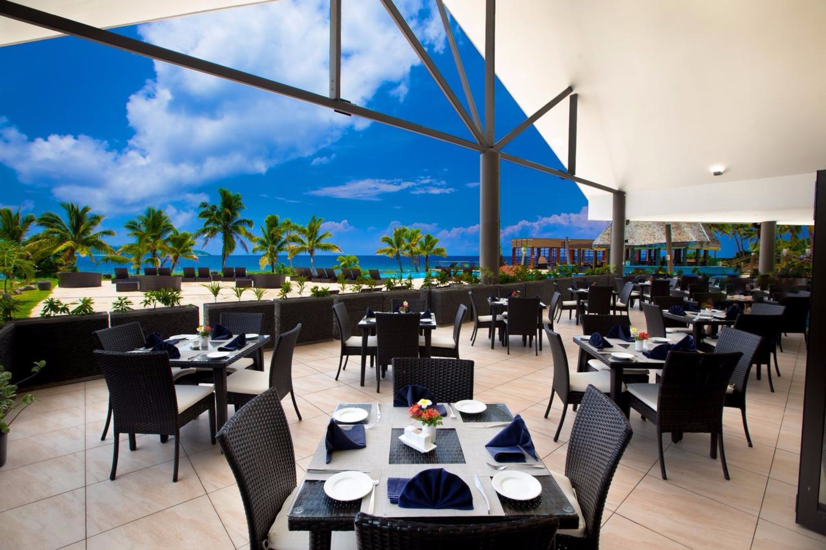 An outdoor dining area with tables and chairs, overlooking a beach with palm trees and the ocean.