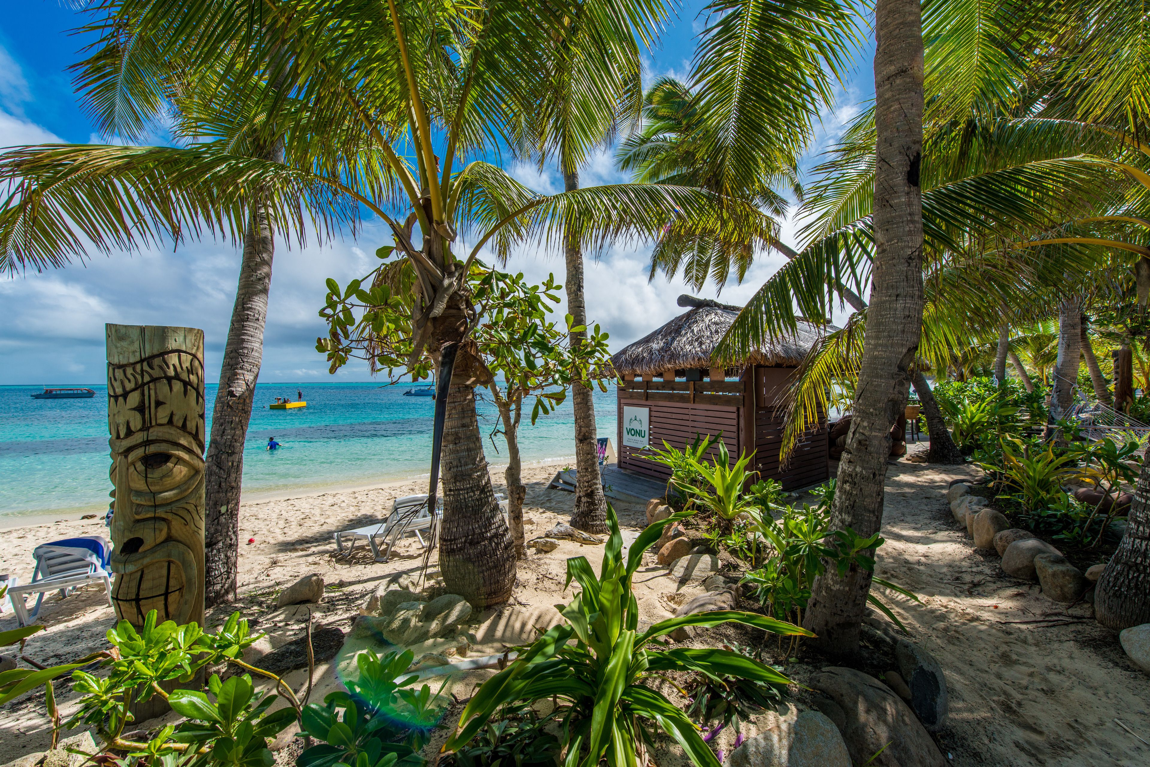 Une plage bordée de palmiers, une hutte au toit de chaume et un poteau en bois orné de sculptures.