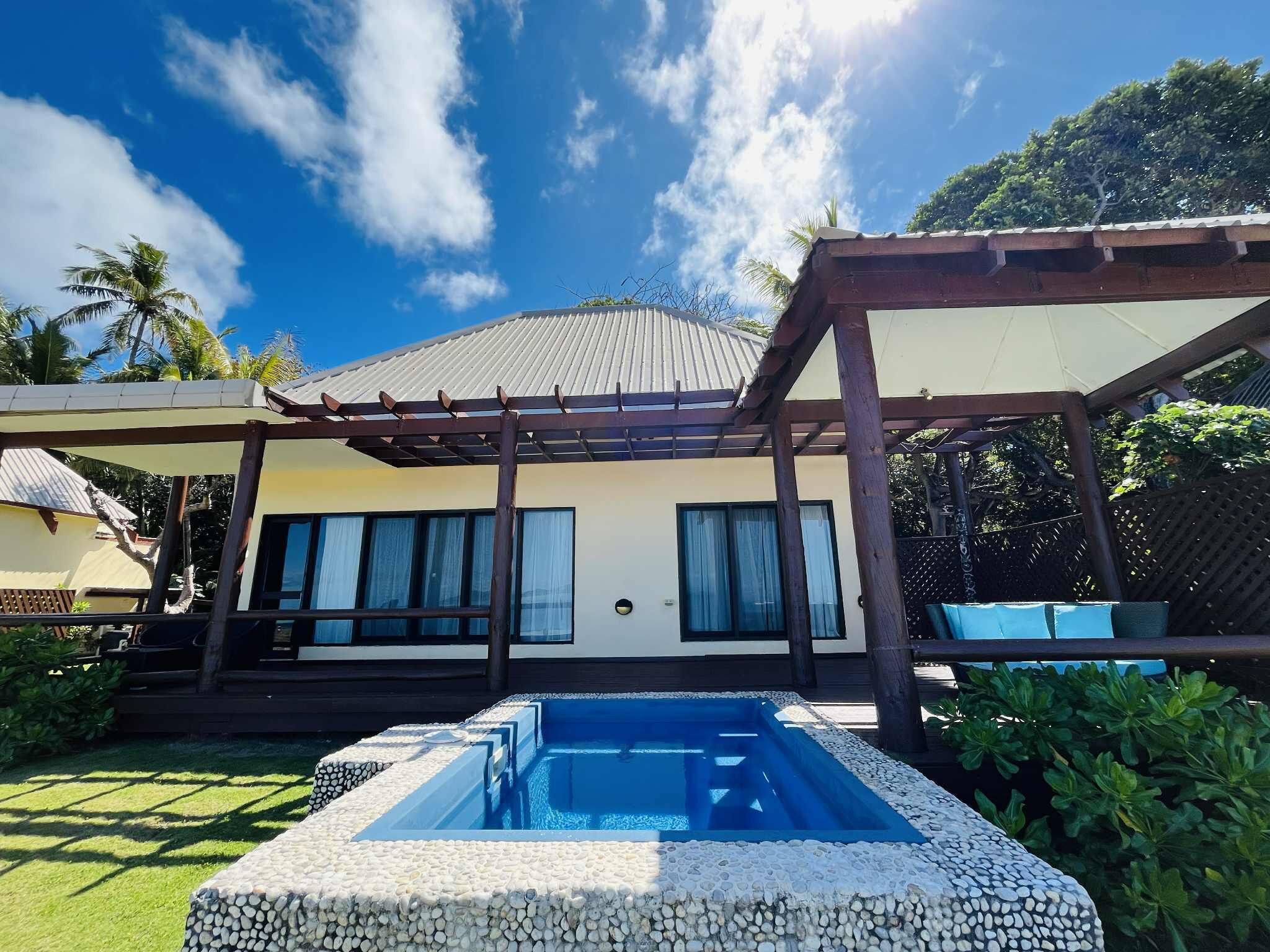A pool with a pebble border, a covered patio area, and a building with large windows and a sloped roof.