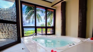 Un baño con una bañera grande, una ventana con vista a palmeras y al océano, y una pared de piedra.