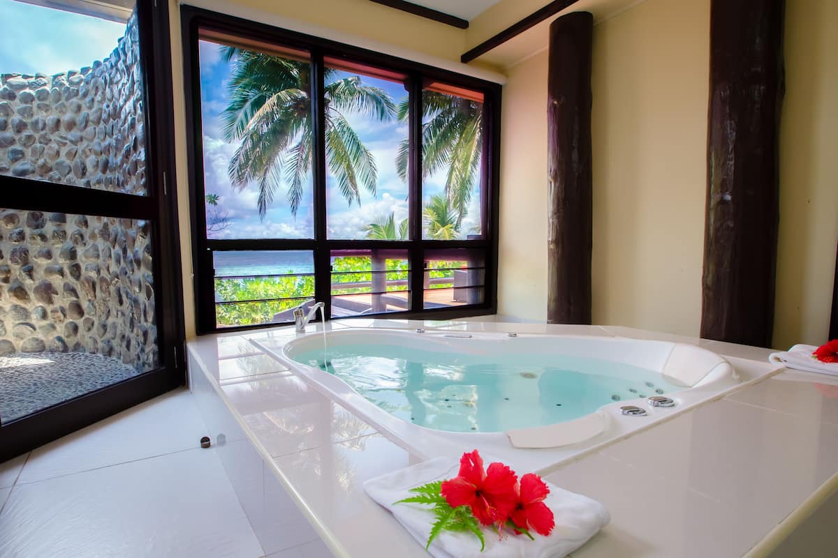 A bathroom with a large bathtub, a window with a view of palm trees and the ocean, and a stone wall.