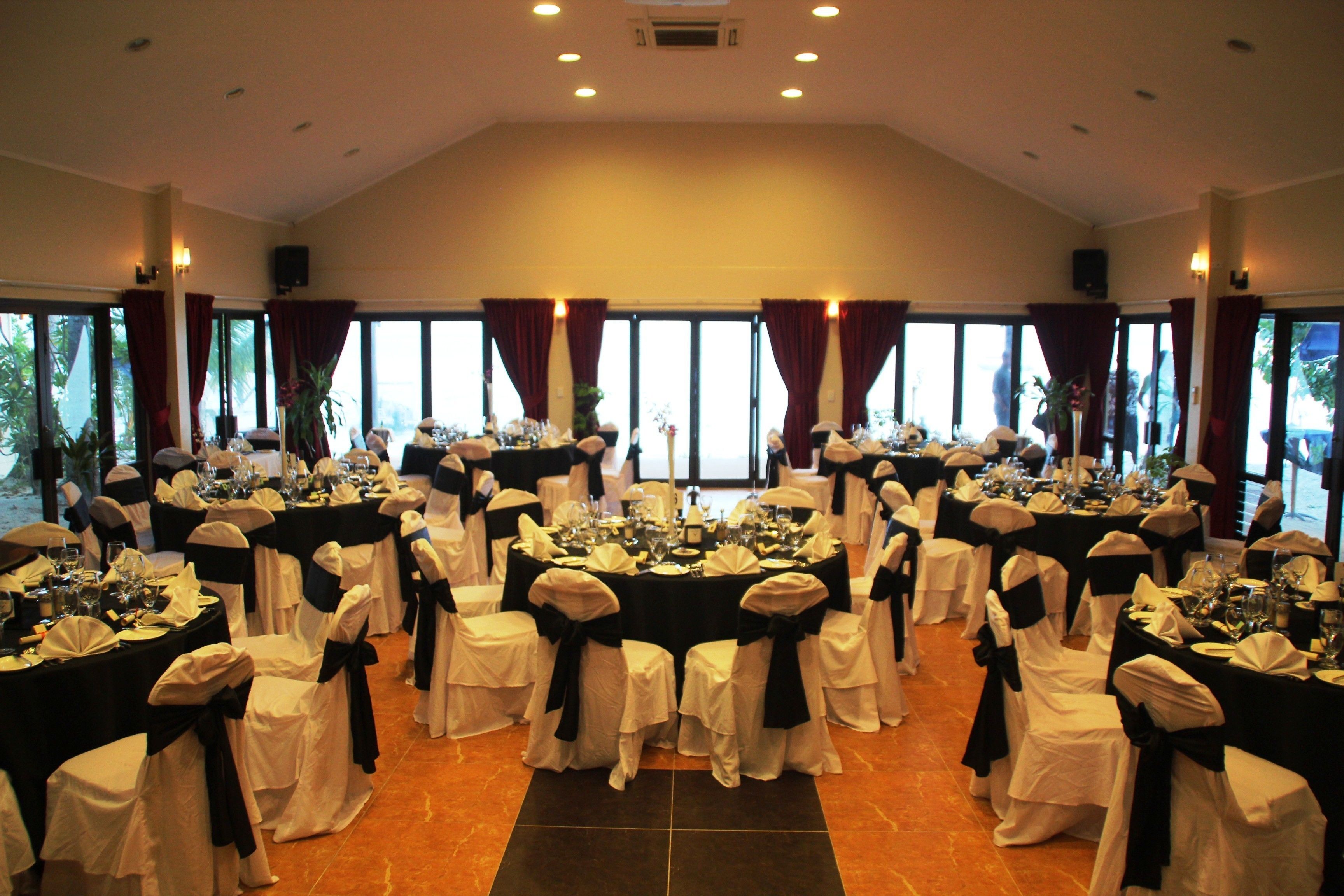 Une salle de banquet avec des tables rondes, des chaises blanches ornées de rubans noirs et de grandes fenêtres avec des rideaux.