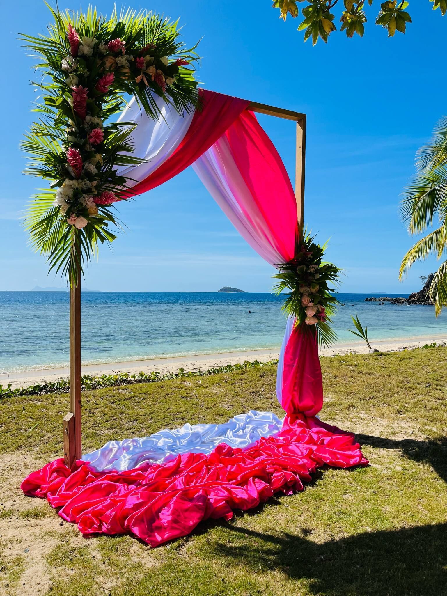 A beach wedding setup with a wooden arch, floral decorations, and a red and white draped fabric.