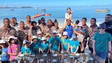 A group of people gathered around a grill with food items on it, with a body of water and boats in the background.