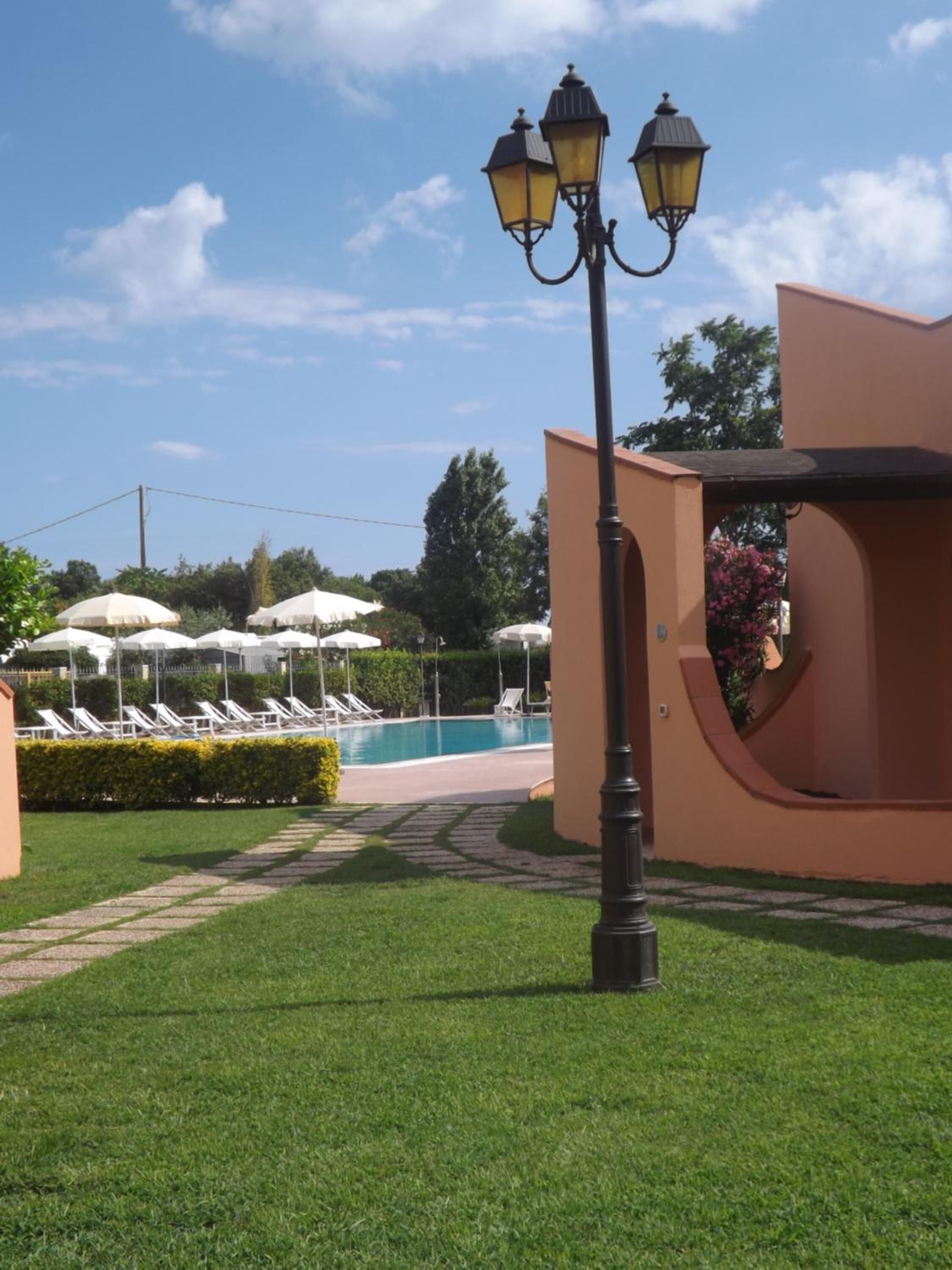 A poolside area with lounge chairs and umbrellas, a lamp post, and a pathway leading to a building.