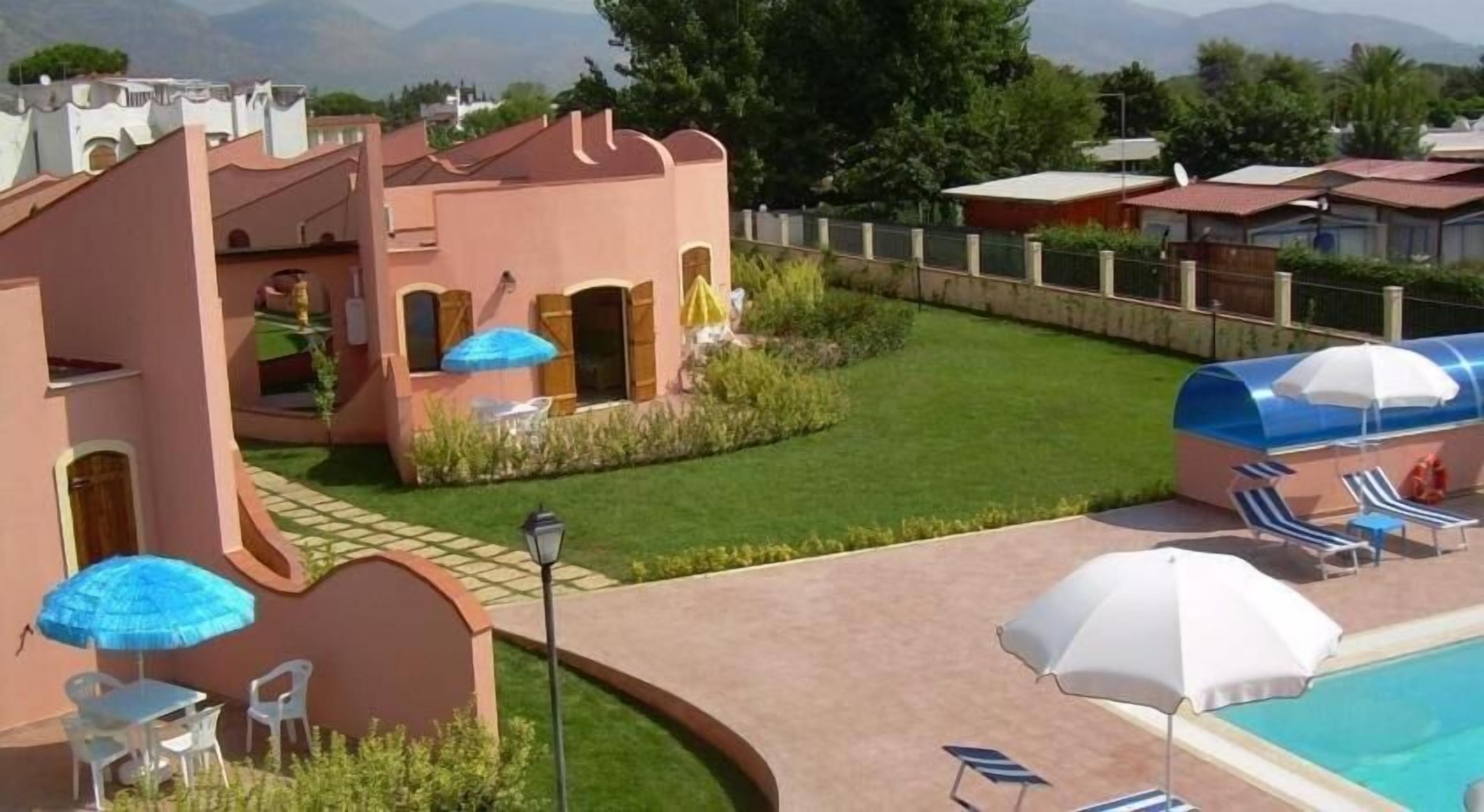 Zona de piscina amb gandules i ombrel·les, un petit jardí i un edifici rosa amb portals arquejats.