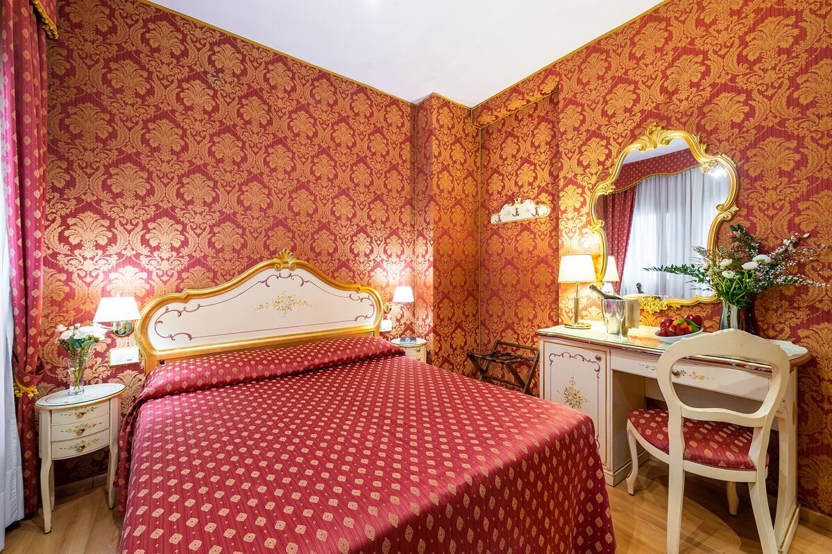 Ein Zimmer mit einem Bett, einem Schreibtisch, einem Stuhl, einem Spiegel und einem Nachttisch mit Lampe und Blumen.