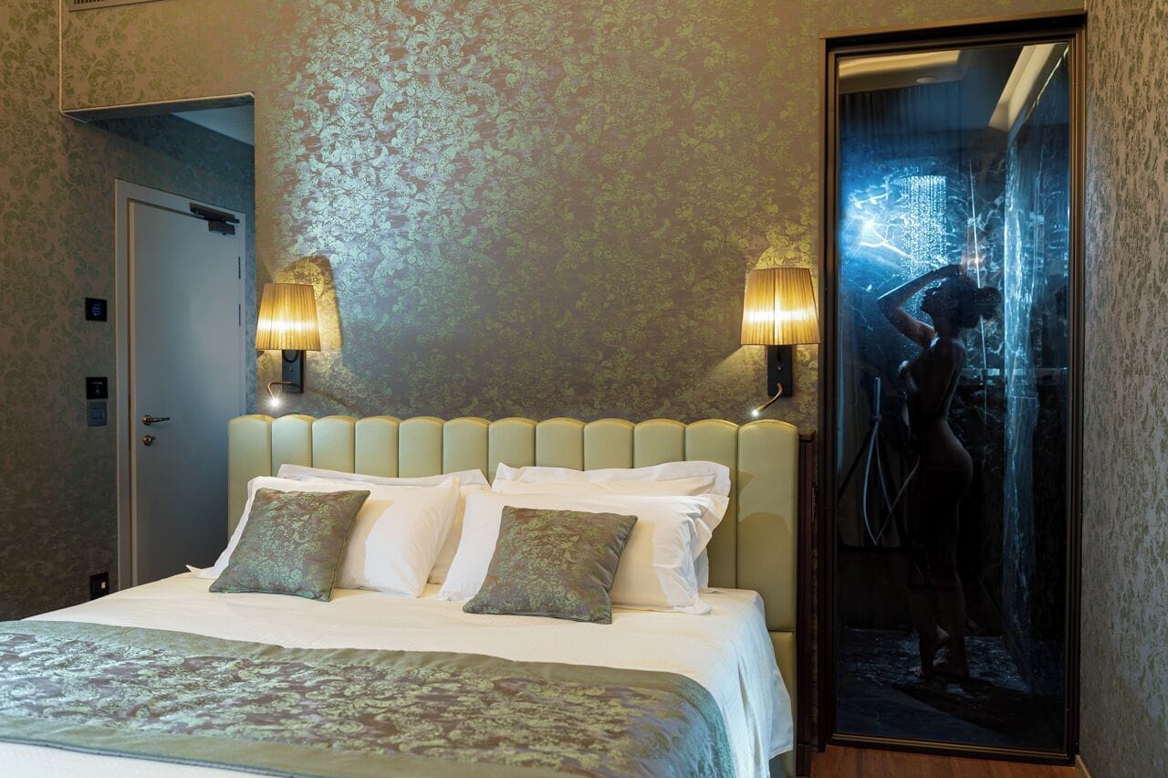 A hotel room with a bed, two wall-mounted lamps, and a large mirror reflecting a shower area.