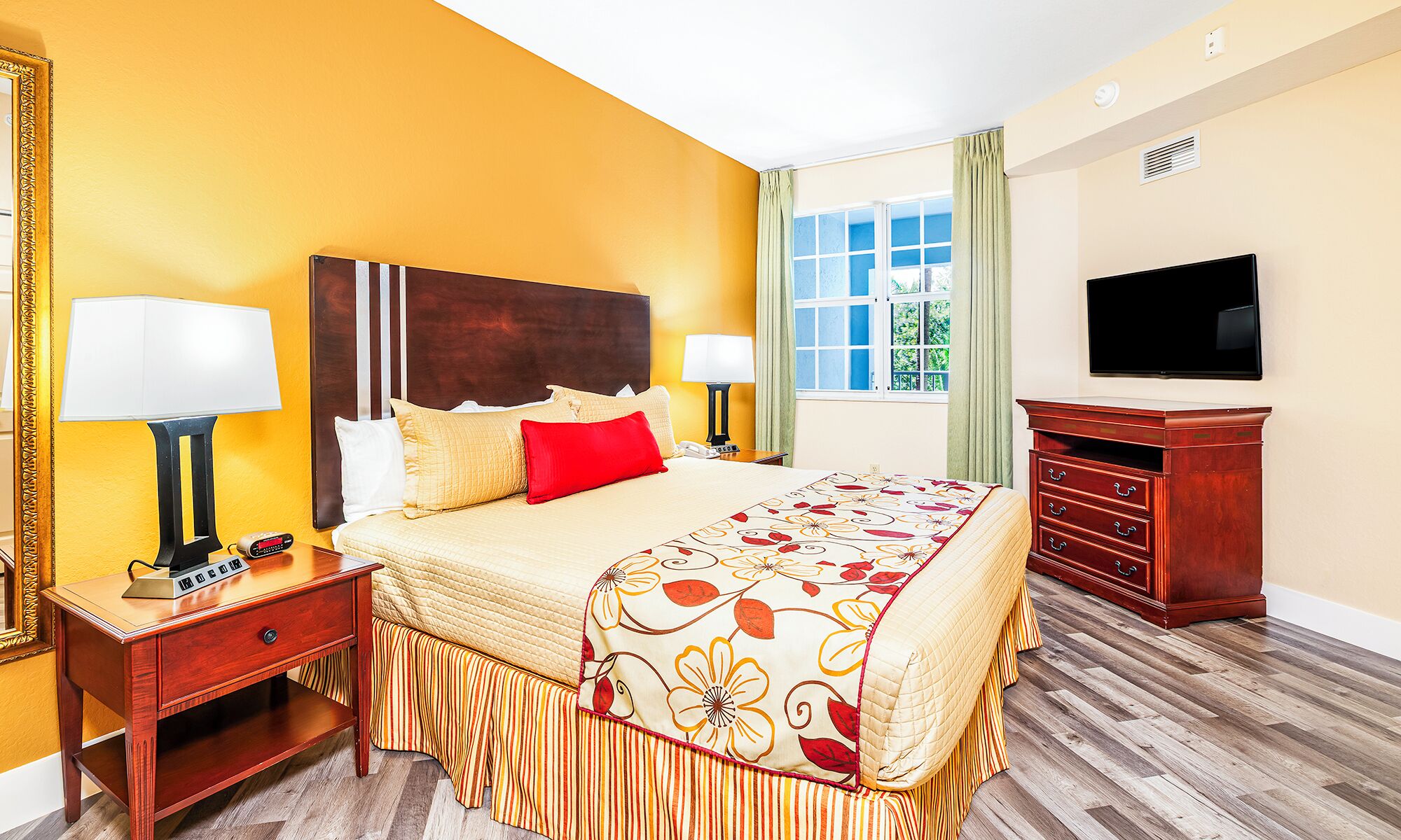 A hotel room with a large bed, two bedside tables with lamps, a wooden dresser, a television on the wall, and a window with curtains.
