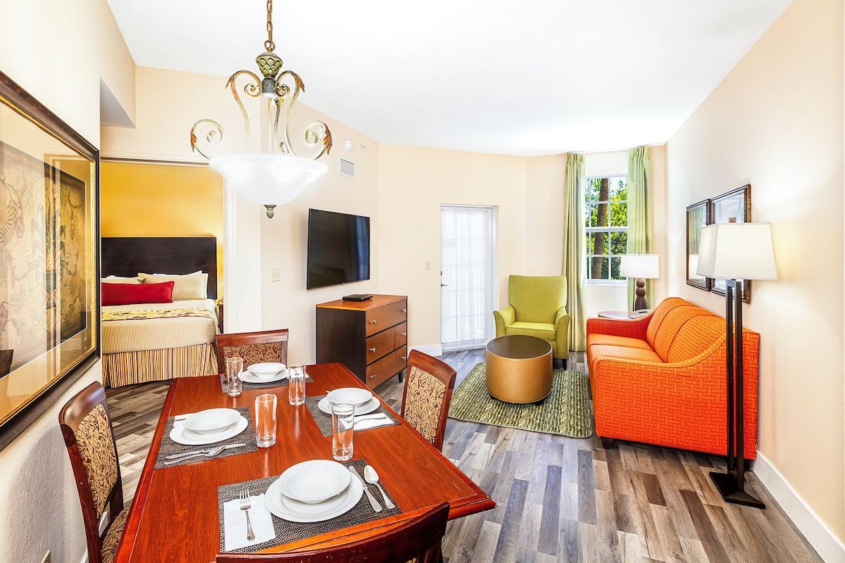 A hotel room with a dining area, a bed with a headboard, a television, a sofa, and a chair.