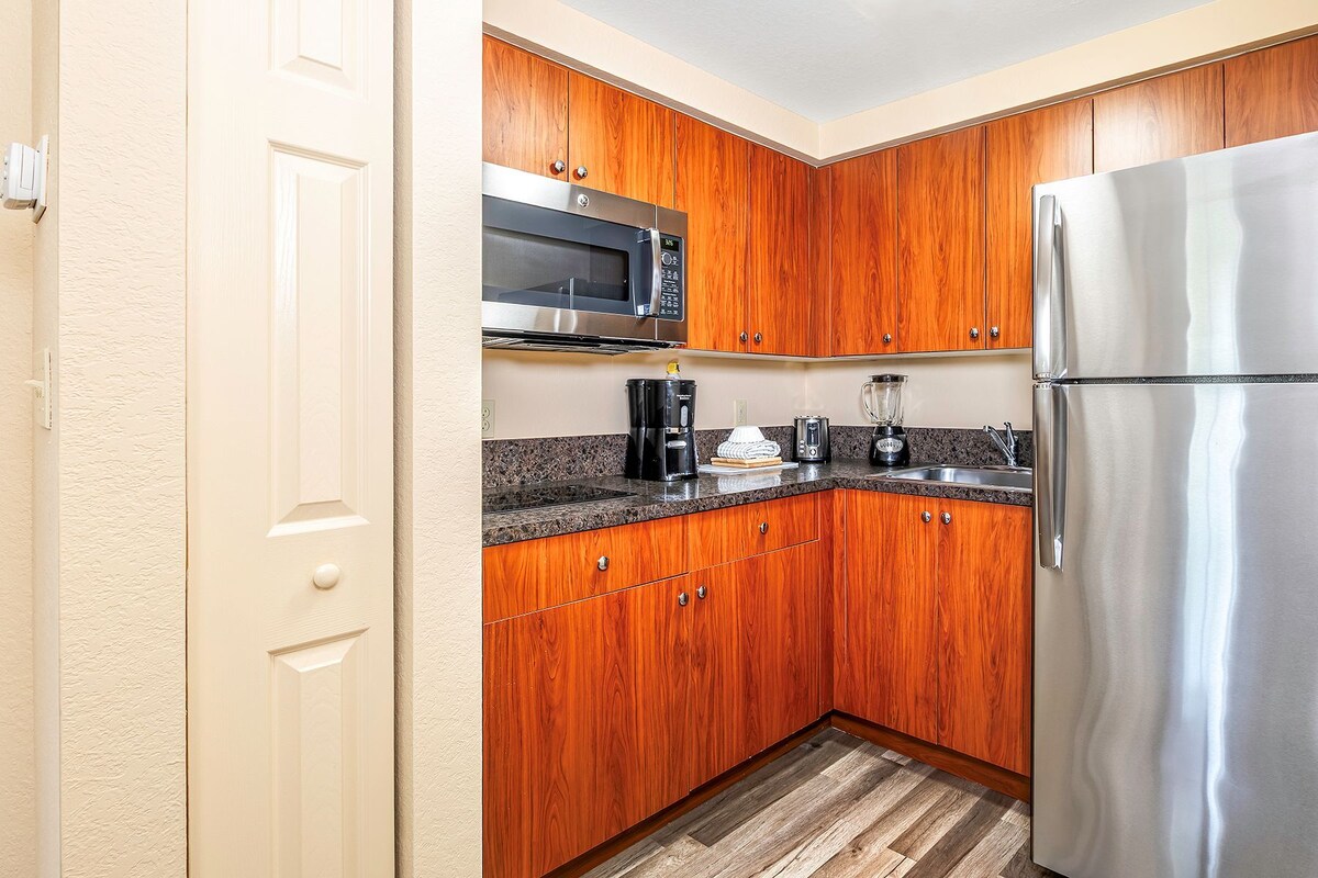 Una cocina compacta con gabinetes de madera, un refrigerador de acero inoxidable, un microondas y una cafetera.