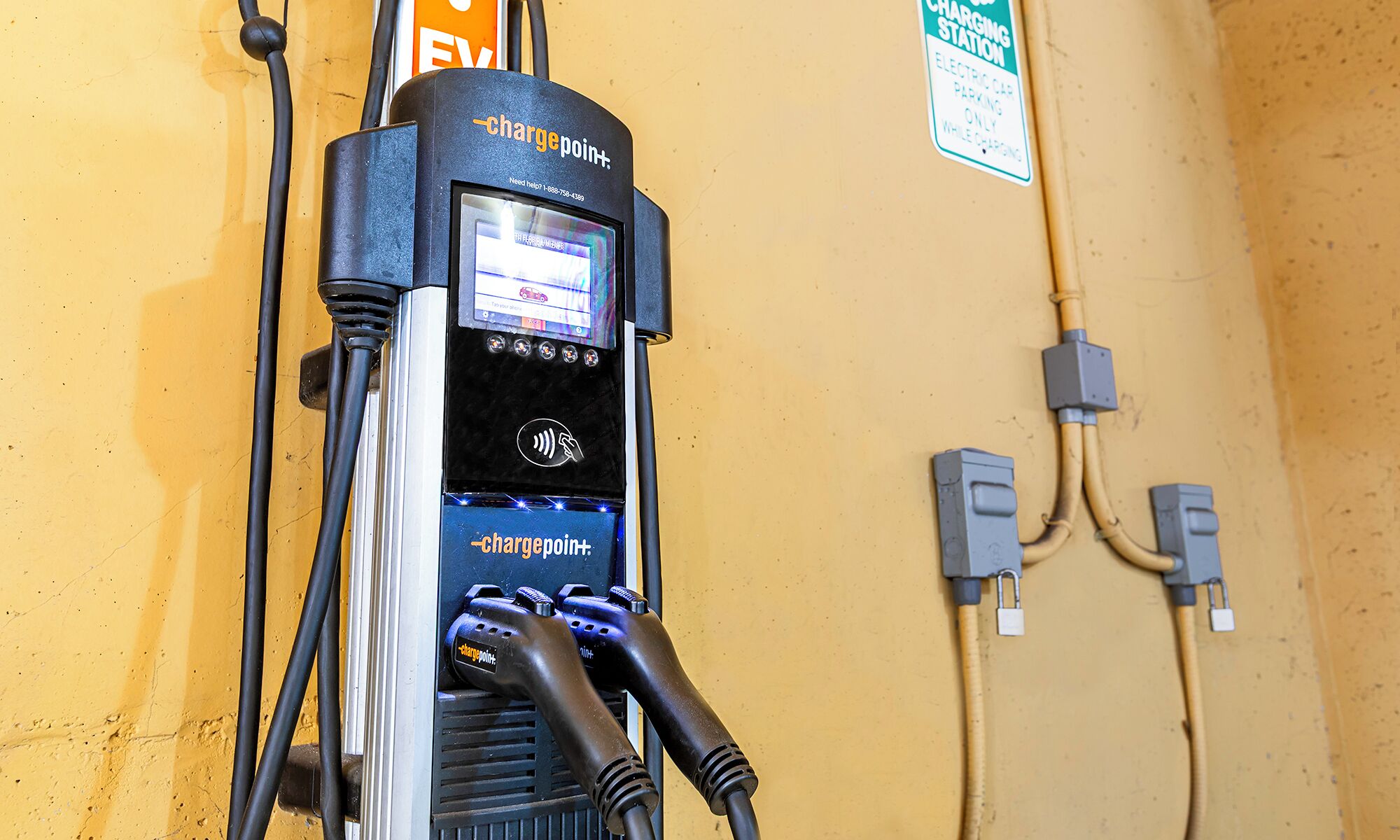 A ChargePoint electric vehicle charging station with a digital display, charging cables, and a wall-mounted sign indicating it is a charging station.