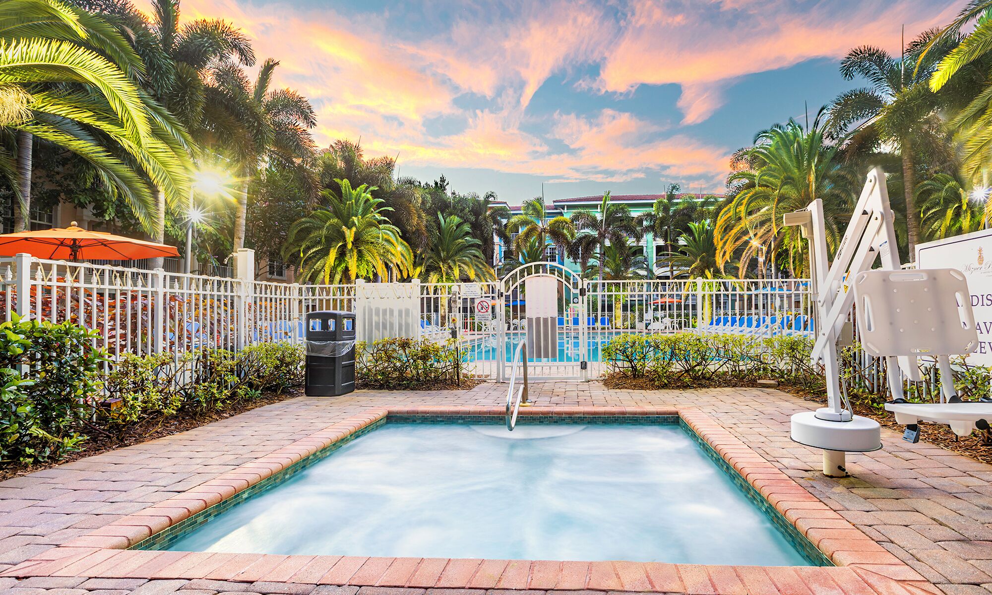 A swimming pool with a tiled edge, surrounded by a white fence, palm trees, and a building with a red roof in the background.
