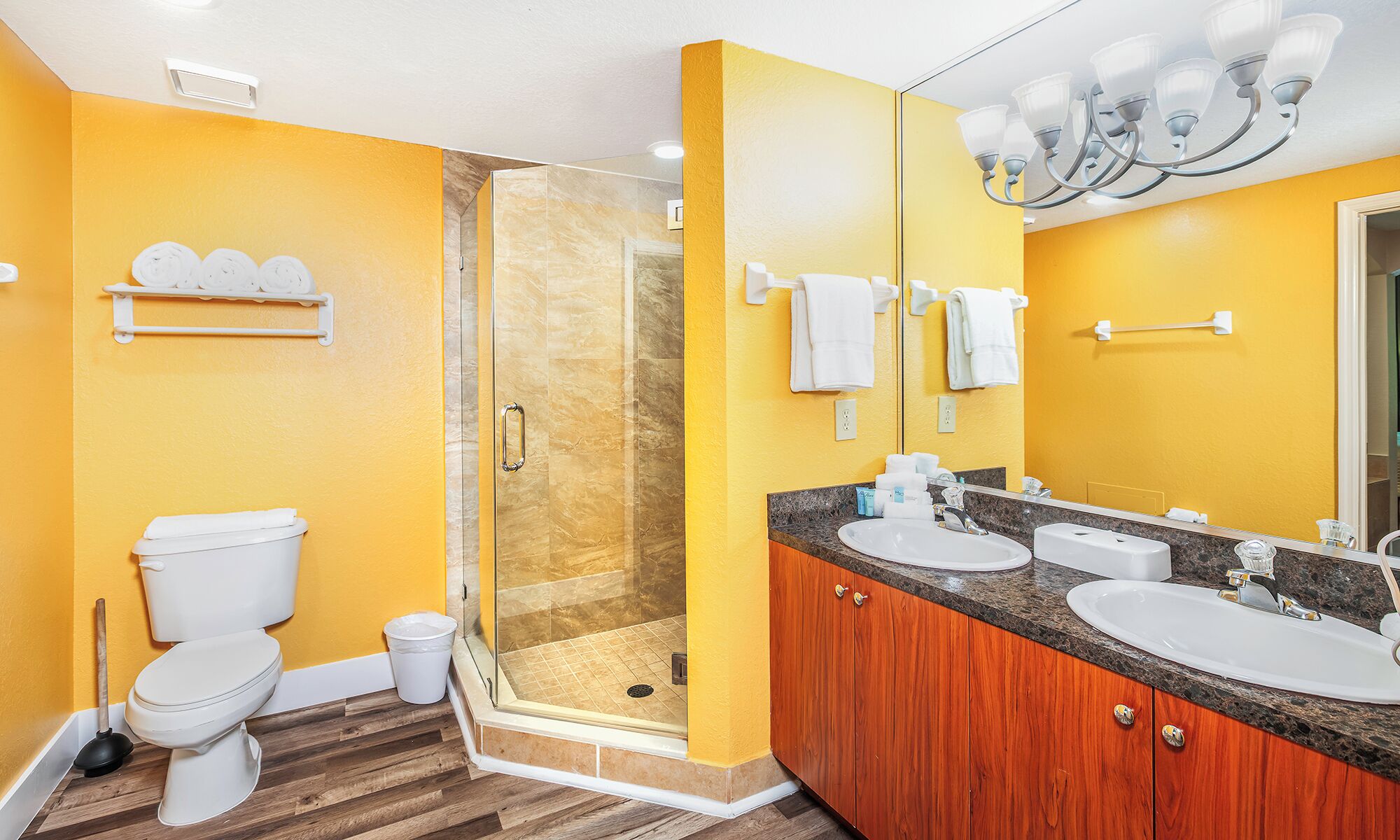 A bathroom with yellow walls, a glass shower enclosure, a white toilet, and a wooden vanity with two sinks.