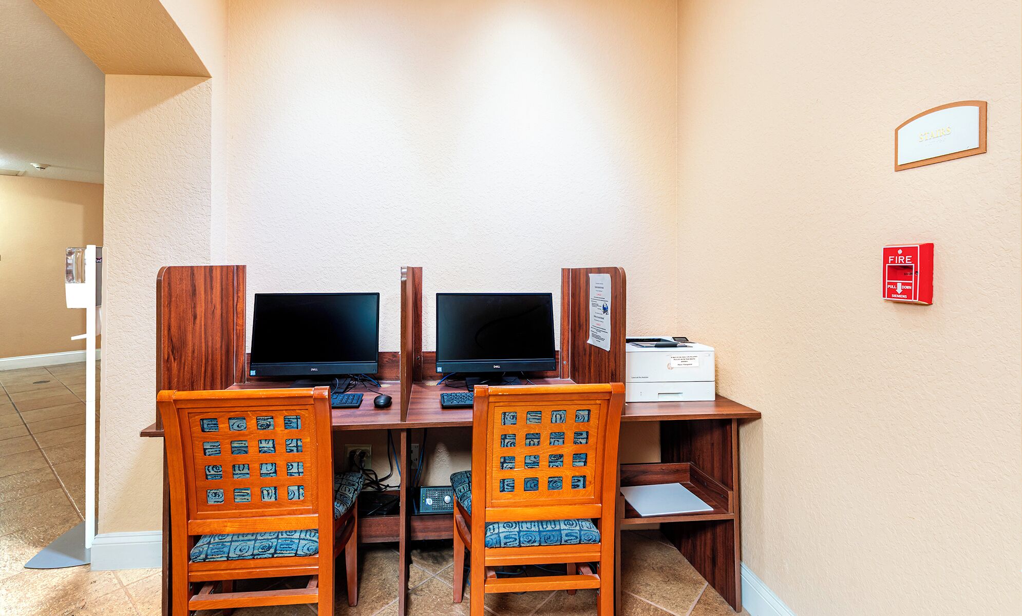 A hotel room with two wooden desks, each with a computer and keyboard, and matching chairs.