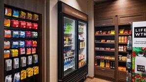 Ett litet kök i hotellrummet med ett kylskåp fyllt med drycker och snacks, en hylla med förpackade livsmedel och en uppsättning flaskor med vatten.