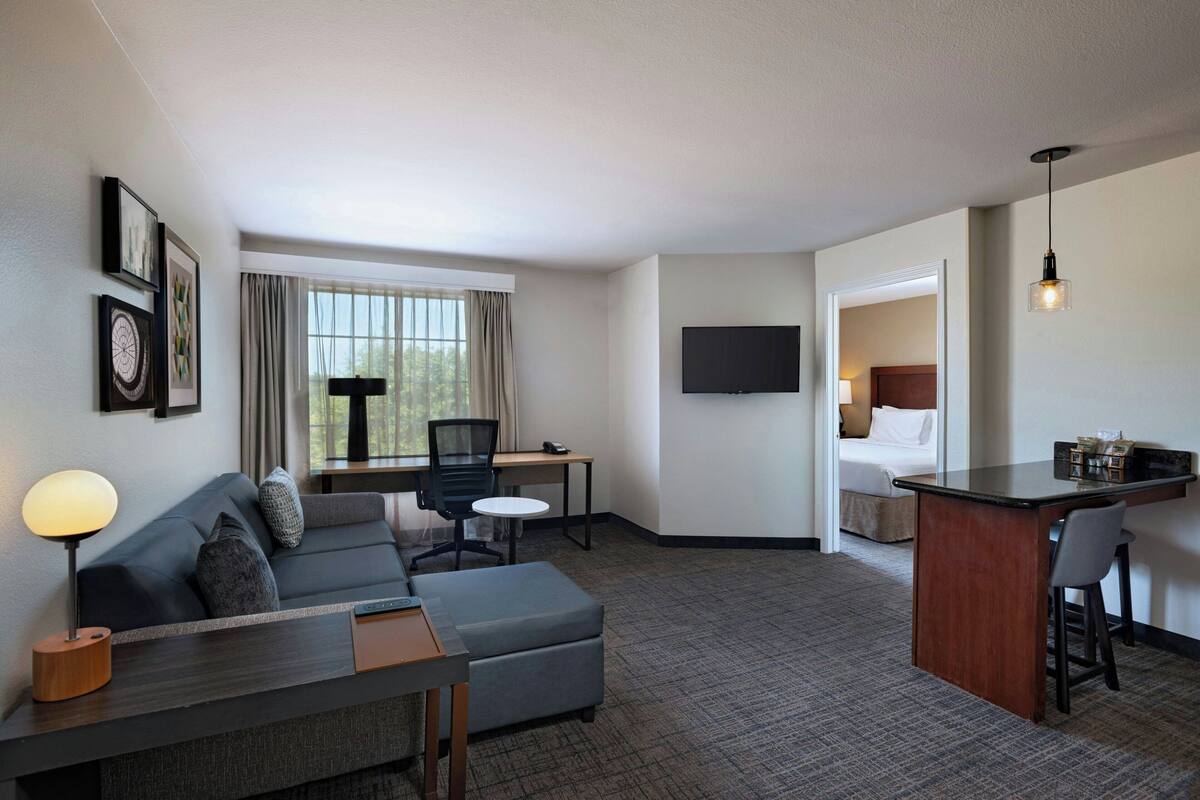 A hotel room with a sofa, ottoman, desk, and a small kitchenette area.
