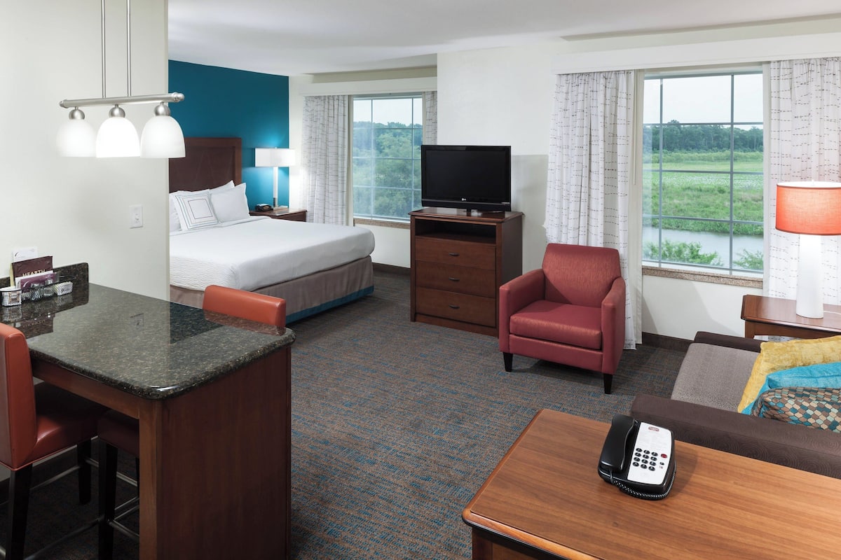 A hotel room with a bed, a television, a desk with a phone, a chair, and a view of a green landscape through the window.