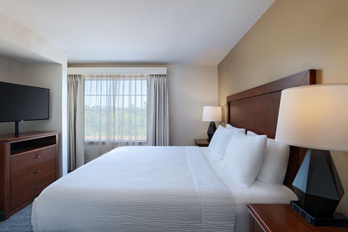 A hotel room with a large bed, a TV on a wooden dresser, a nightstand with a lamp, and a window with curtains.