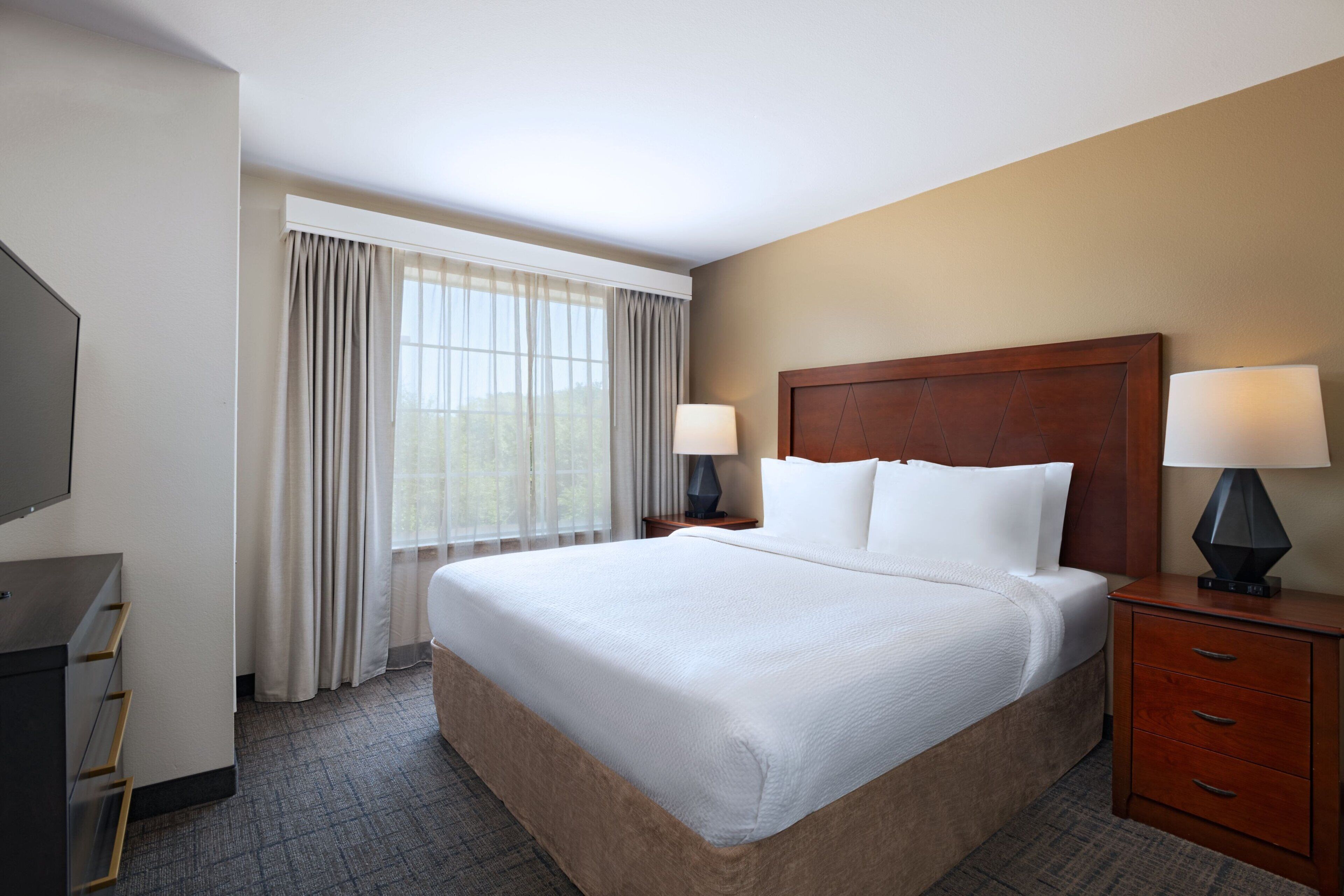 A hotel room with a large bed, two bedside tables with lamps, a TV, and a window with curtains.