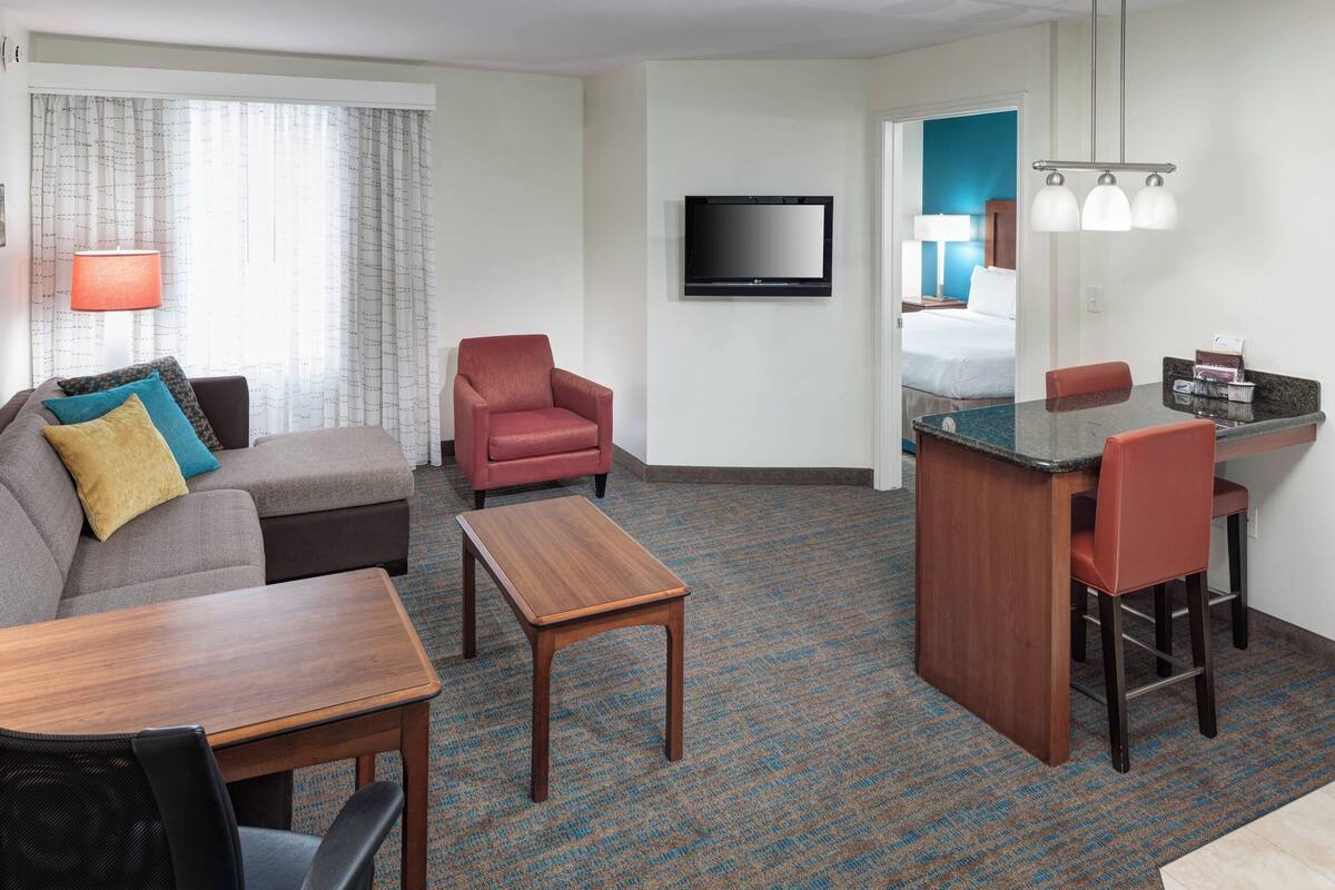A hotel room with a sofa, armchair, small table, and a kitchenette area with a TV and a mirror.