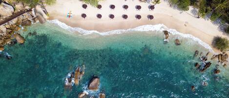Vista aérea de una playa con agua turquesa cristalina, una orilla arenosa y varias sombrillas y tumbonas dispuestas a lo largo de la playa.