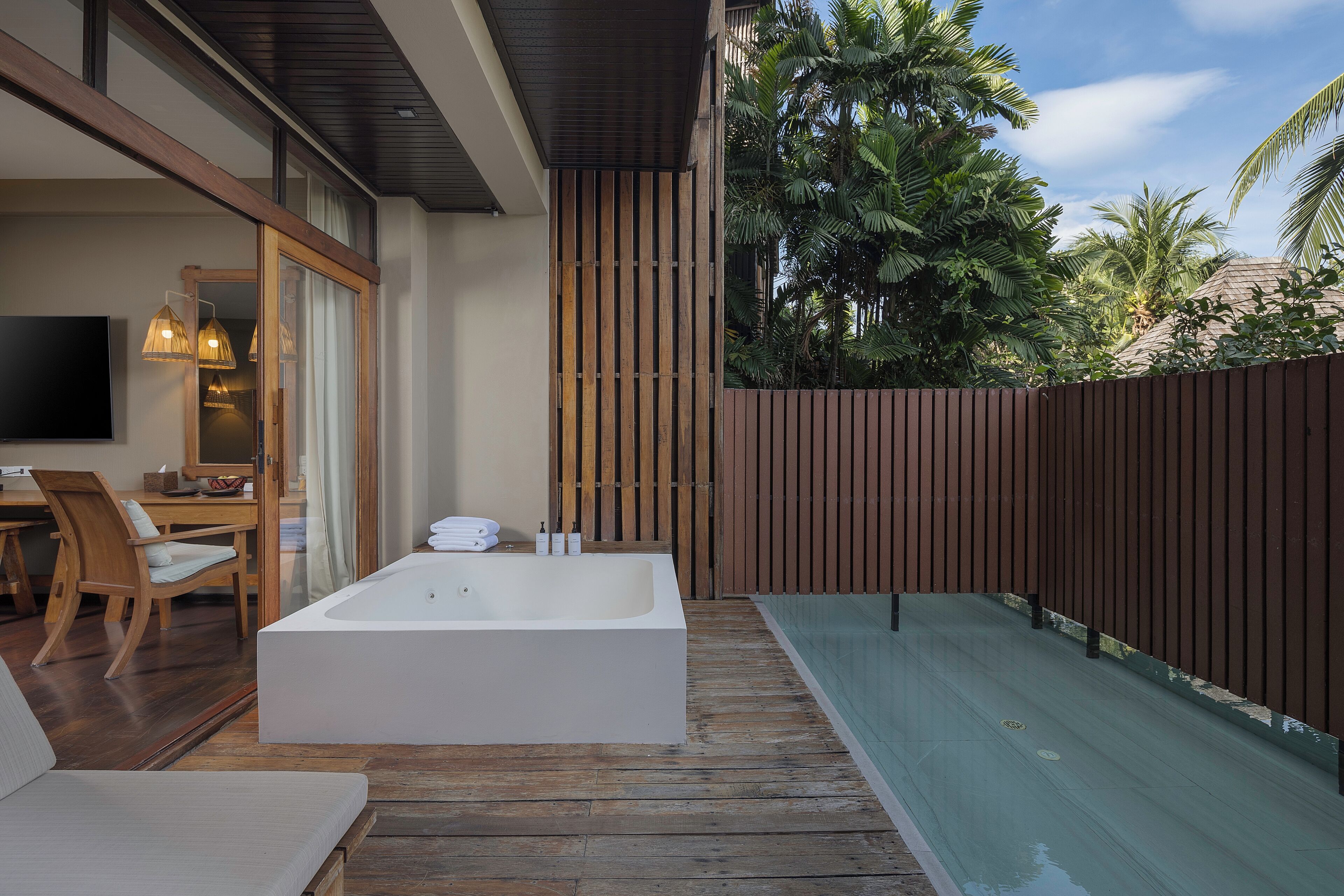 A modern outdoor bathroom with a wooden deck, a large bathtub, and a pool area surrounded by a wooden fence and tropical plants.