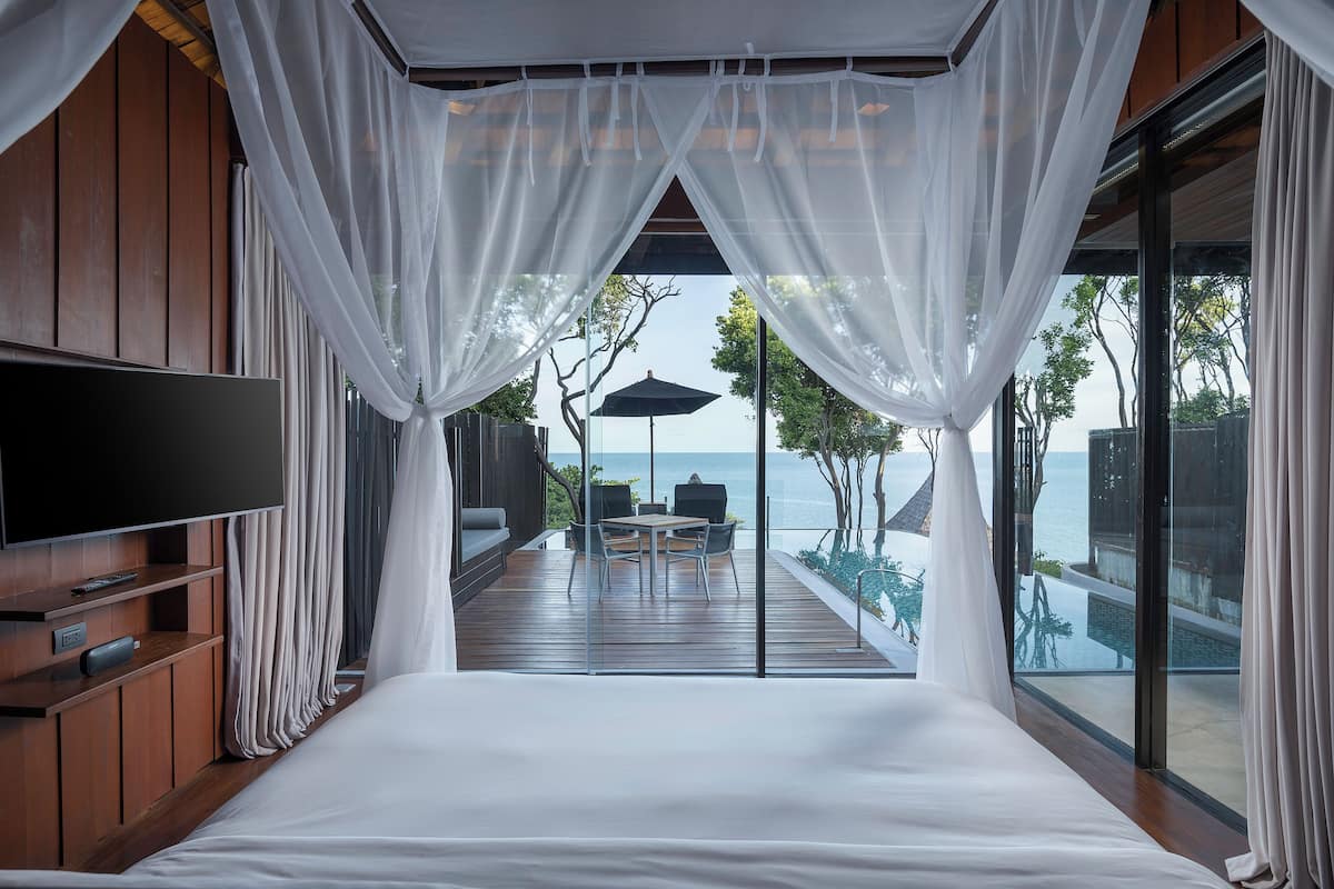 A bedroom with a large bed, a TV, and a view of the sea through sheer curtains.
