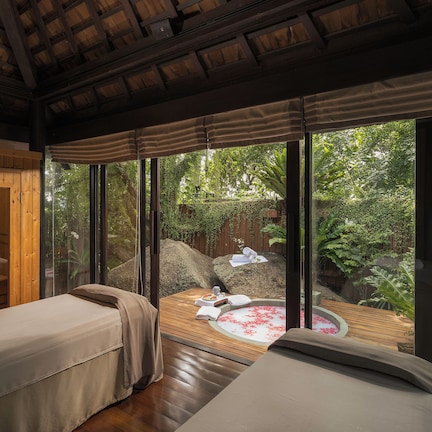 A room with two beds, a wooden ceiling, large windows, a hot tub with rose petals, and a view of a garden.