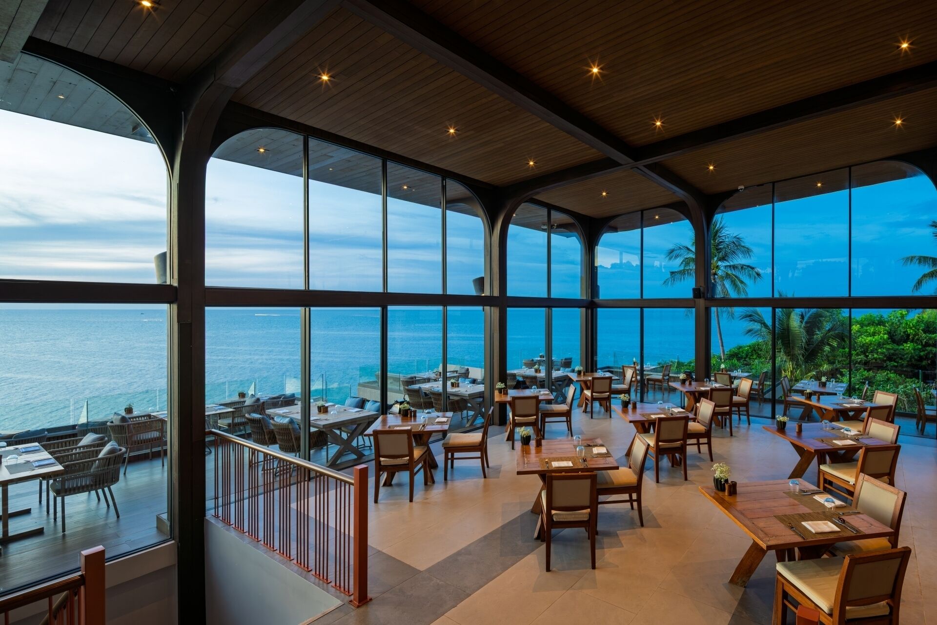 A restaurant with large windows offering ocean views, wooden tables and chairs, and a modern interior design.