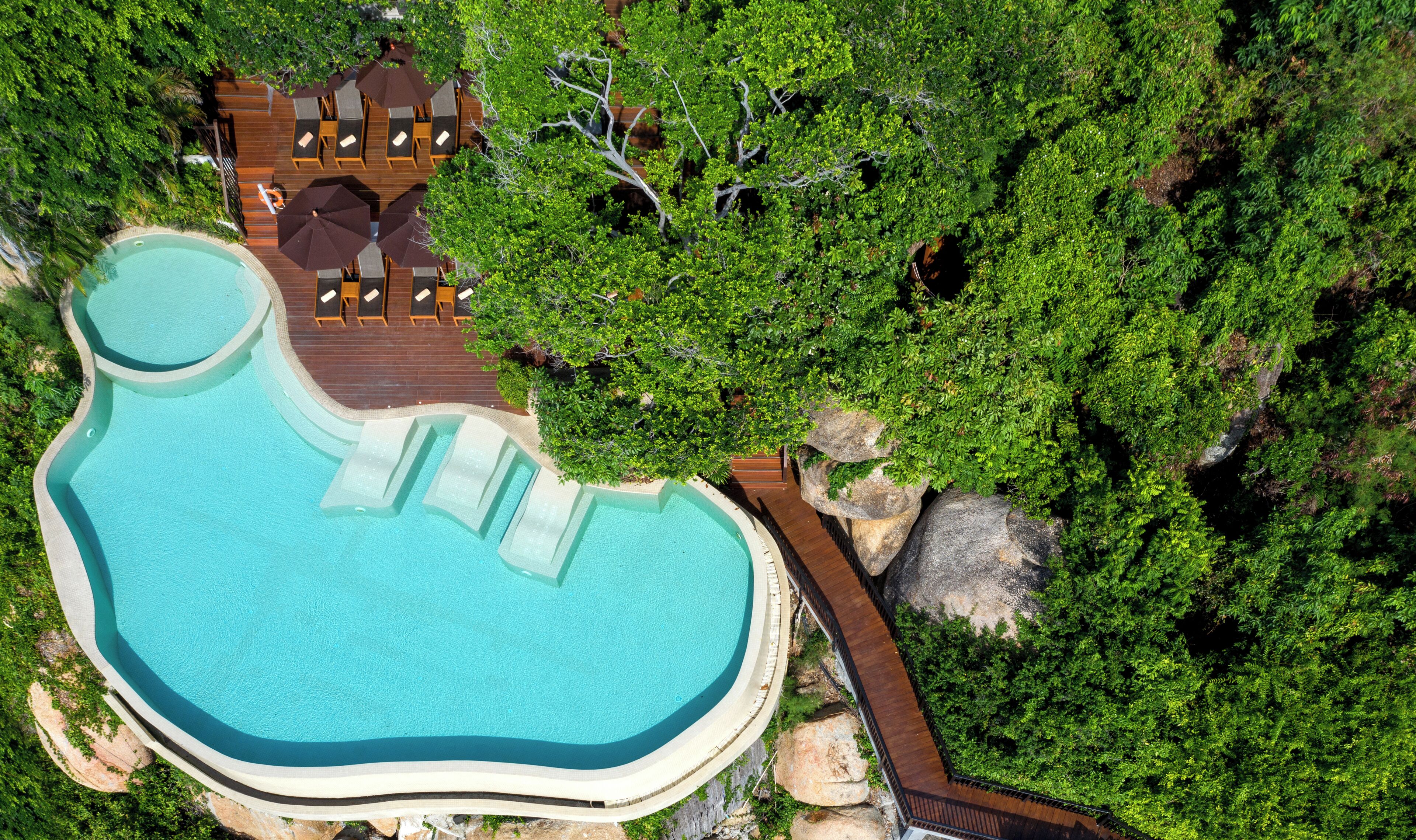 Vista aérea de un complejo turístico con una piscina grande y de forma peculiar, rodeada de exuberante vegetación y una terraza de madera con sillones de descanso.