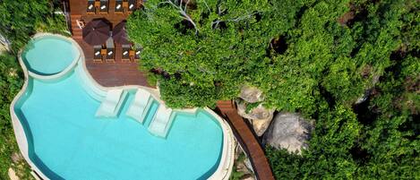 Vista aérea de un complejo turístico con una piscina grande y de forma peculiar, rodeada de exuberante vegetación y una terraza de madera con sillones de descanso.