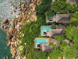 An aerial view of a resort with three separate pools, surrounded by lush greenery and rocky terrain.