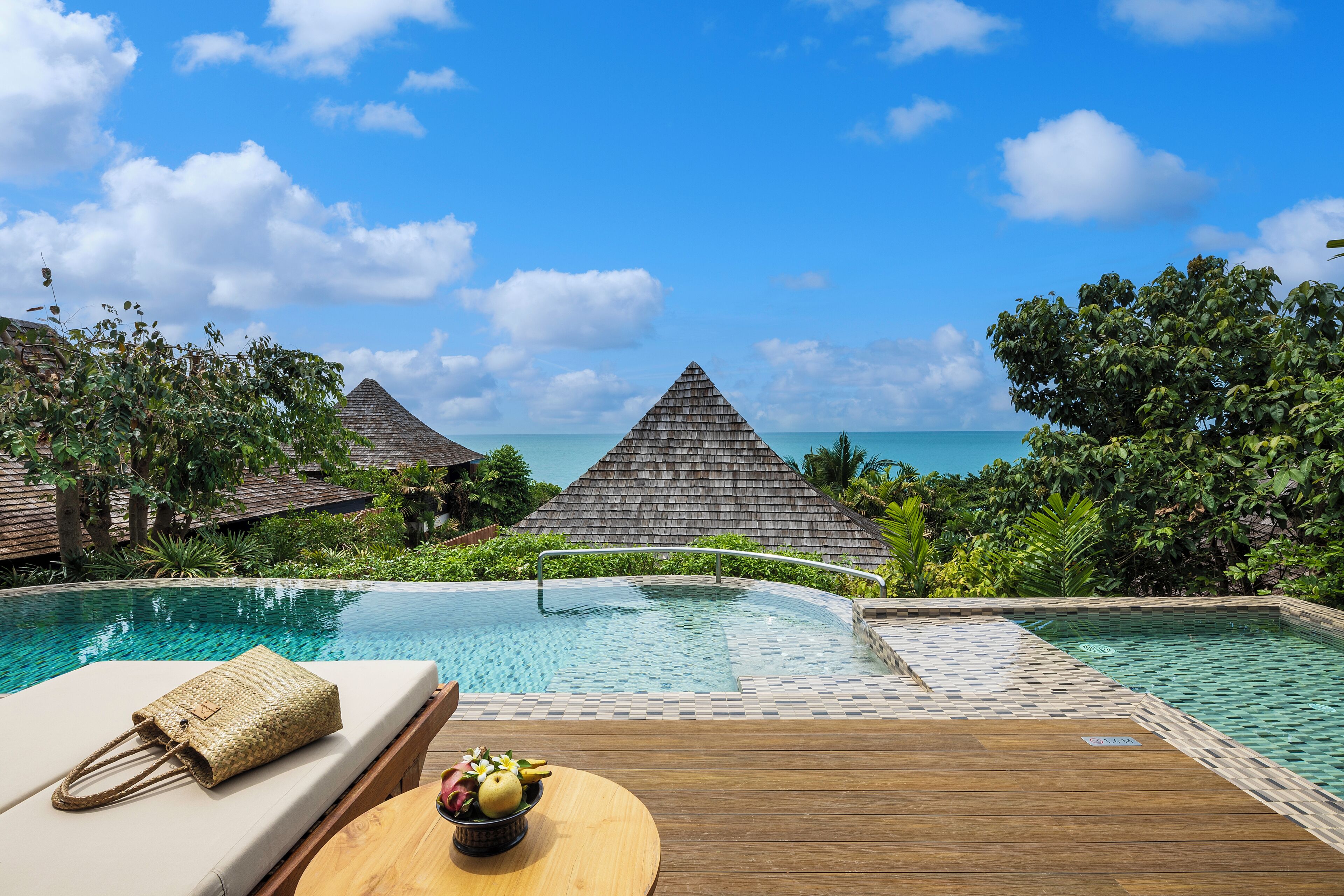 A poolside area with a lounge chair, a wooden deck, and a view of the ocean and resort buildings.