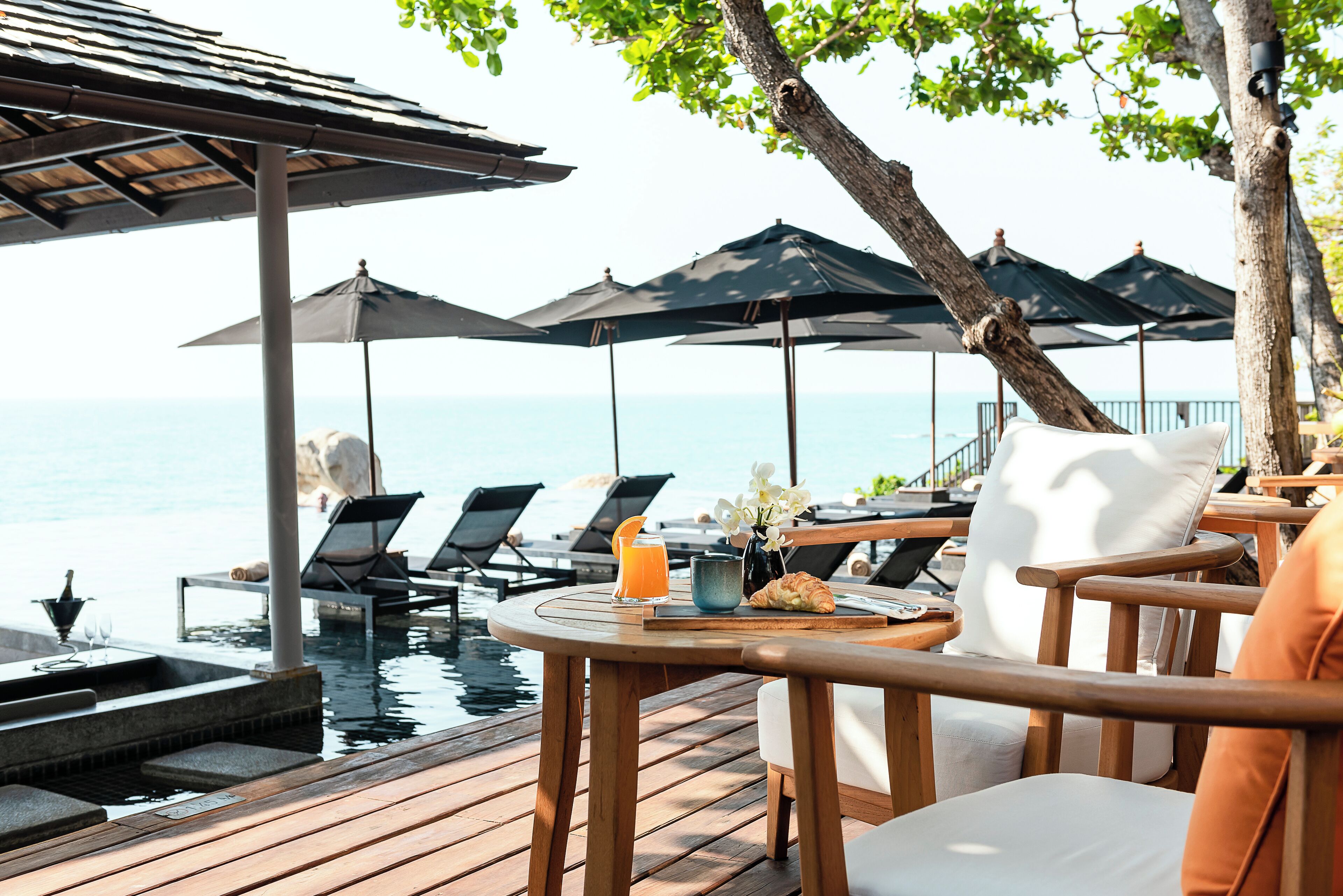 A wooden deck with a pool, lounge chairs, umbrellas, and a table with a drink and pastries.