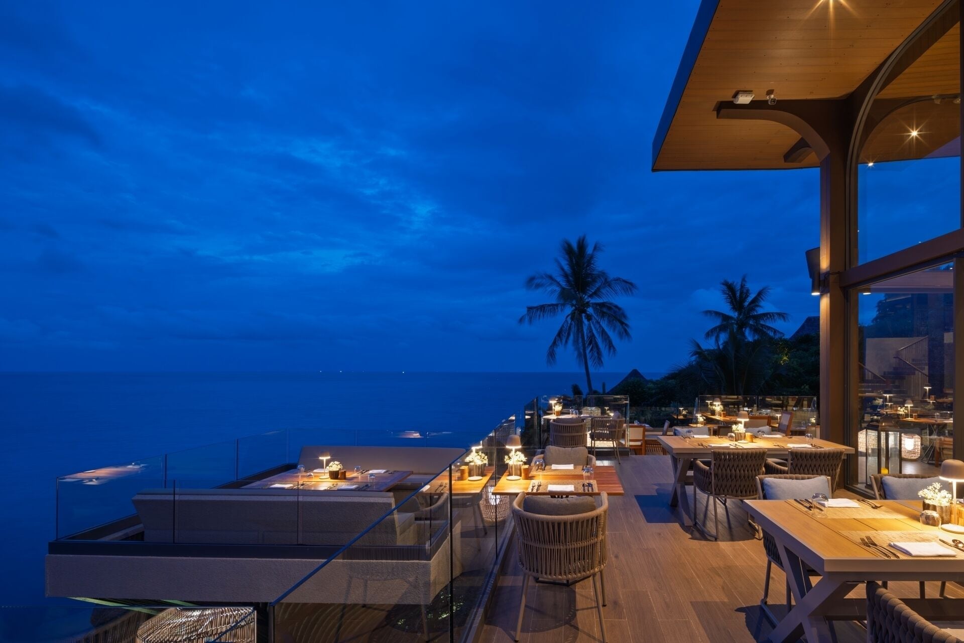 A rooftop dining area with tables and chairs, overlooking the ocean and palm trees.