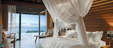 A bedroom with a large bed, a wooden headboard, a nightstand, a bedside table, and a view of the ocean through large windows.