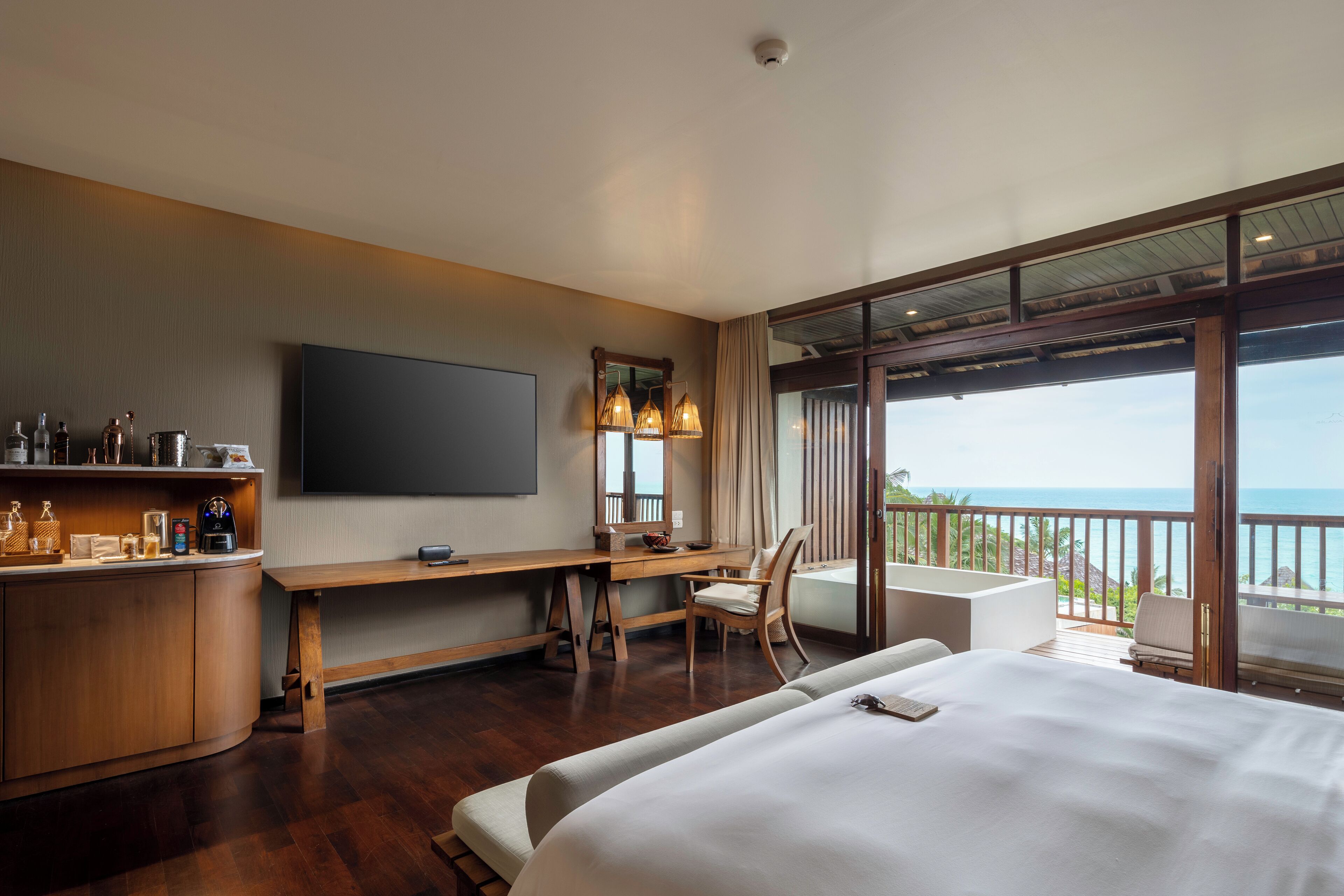 A hotel room with a large bed, a desk with a chair, a TV, and a balcony with a view of the ocean.