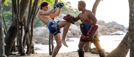 Two people practicing martial arts on a mat outdoors, surrounded by trees and rocks.